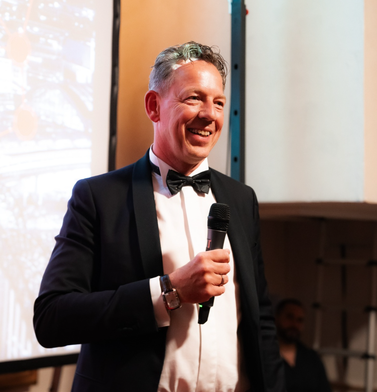 A man in a tuxedo holding a microphone and smiling at an indoor event.