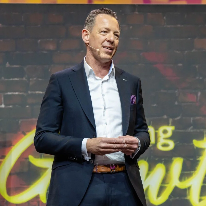 A man in a dark suit and white shirt speaking on stage with a brick wall background and yellow neon text.