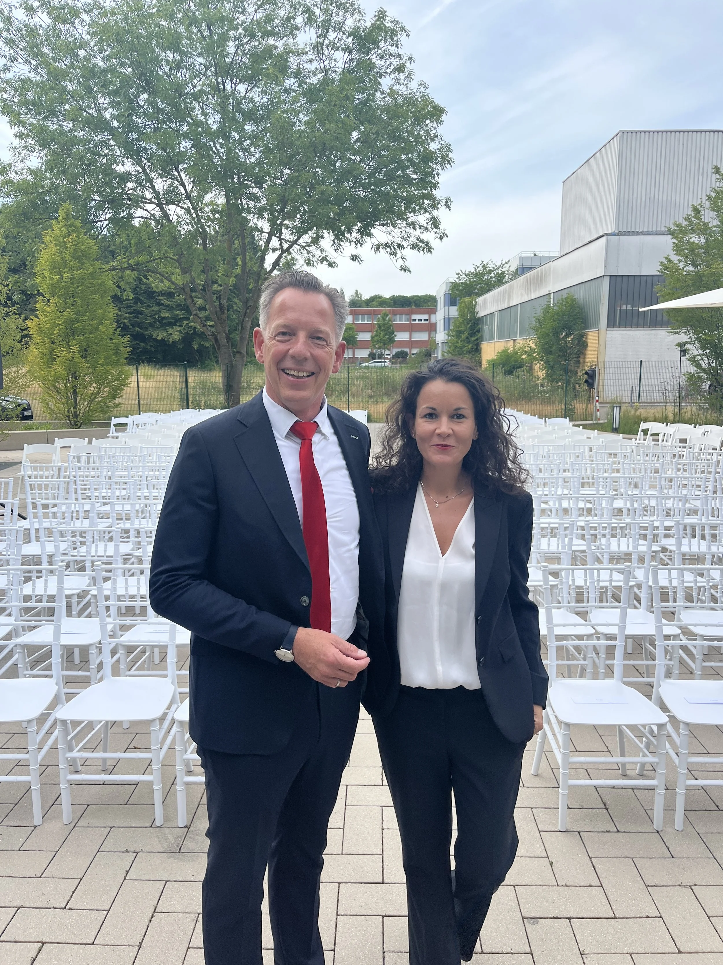 A man and a woman standing outdoors in formal business attire, smiling, with many empty white chairs arranged behind them, trees, a fence, and modern buildings in the background.