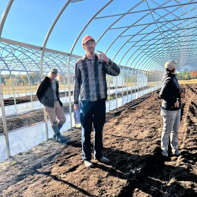 Three people standing inside a large greenhouse built with curved metal arches. The ground is freshly tilled soil, and one person in the center is talking, holding a clipboard and pointing at his face, while the others listen.