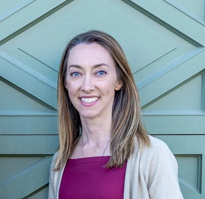 A young woman with shoulder-length light brown hair wearing a purple top and beige cardigan, smiling in front of a green wooden wall.