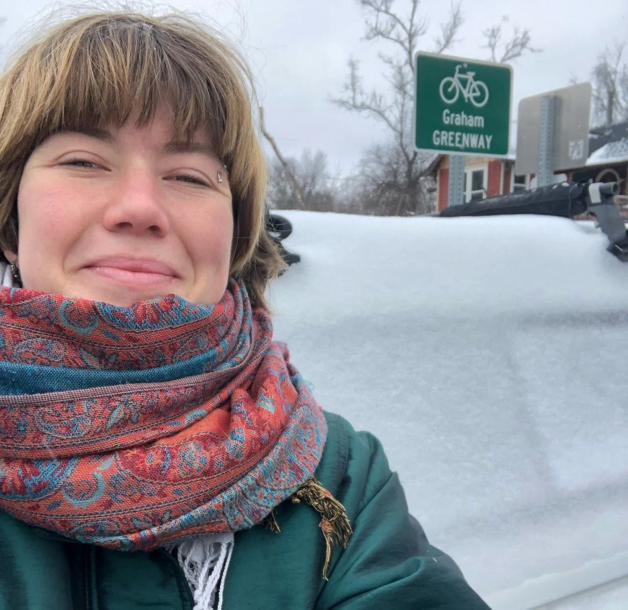 A woman taking a selfie outdoors in a snowy environment, with a sign for Graham Greenway in the background.