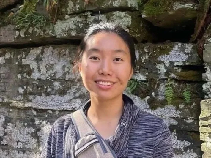 A young woman with dark hair tied back, smiling, standing outdoors in front of a rocky wall with some ferns.