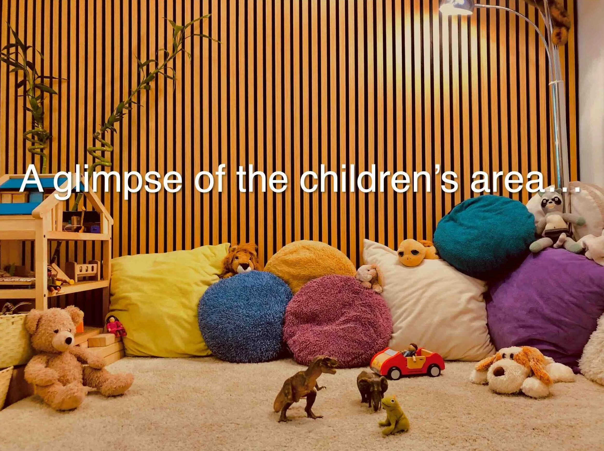 Children's play area with stuffed animals, pillows, and toy dinosaurs on a carpeted floor, against a wooden slat wall.