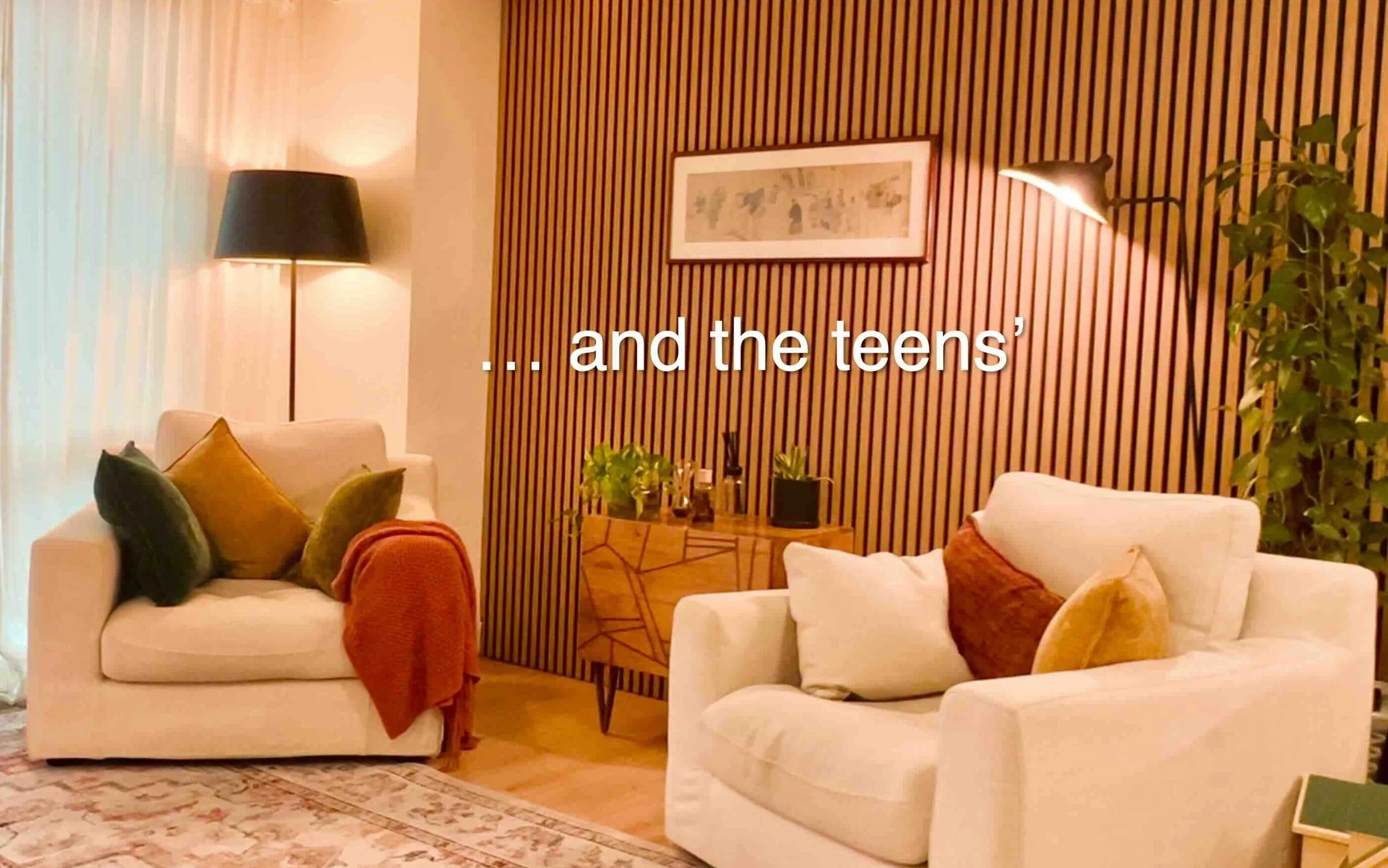 Living room with white sofas and colorful cushions, a wooden side table with plants, a textured rug, and a wall with wooden slats and framed artwork, illuminated by floor and wall lamps.