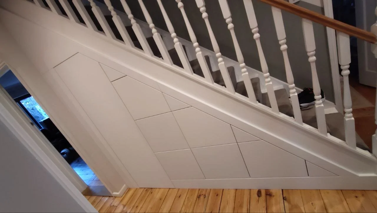 Interior view of a staircase with a custom built storage cabinet underneath, featuring white paneling with geometric patterns, wooden floor, and a railing with white balusters and a wooden handrail.