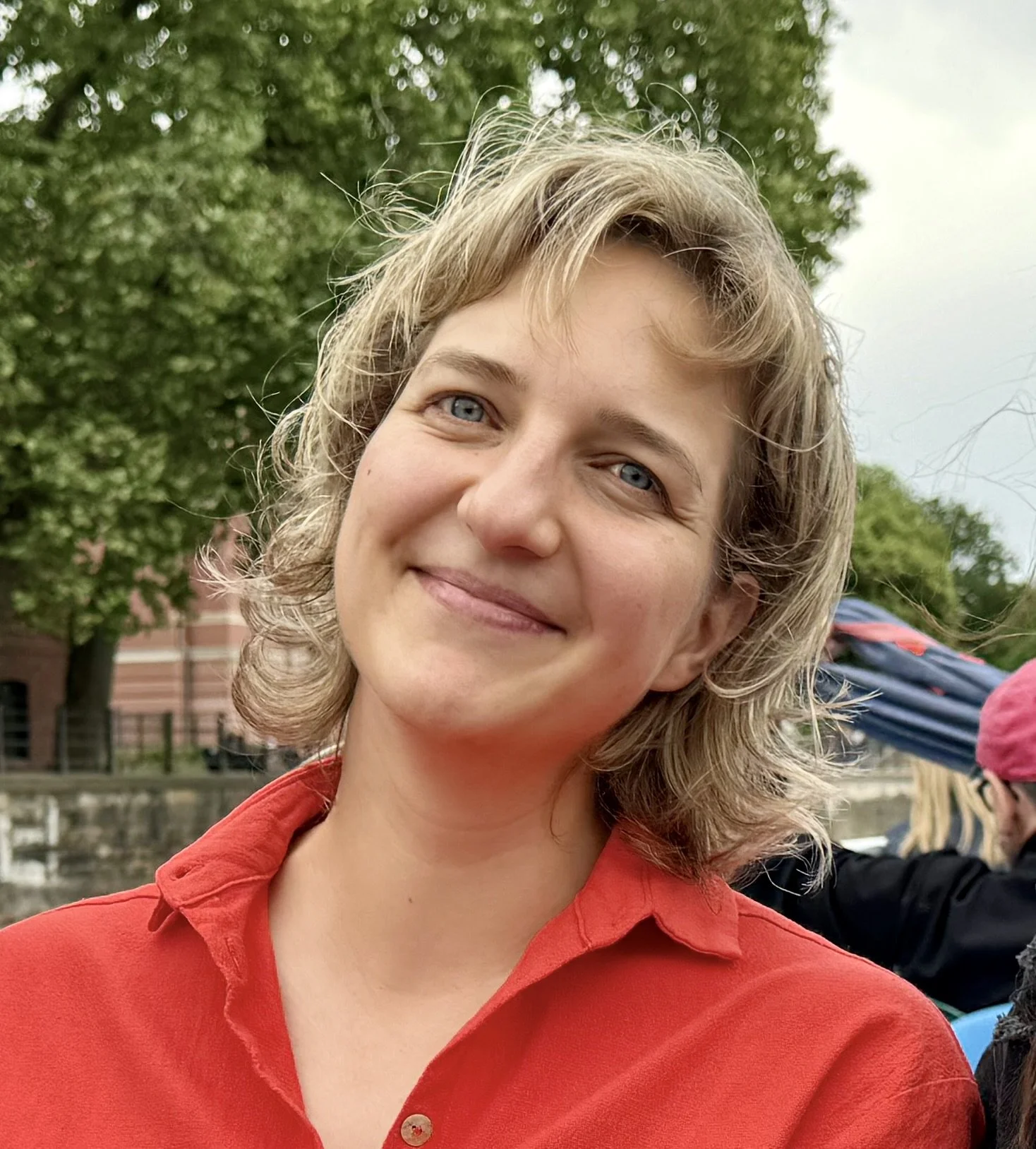 Lächelnde junge Frau mit blauen Augen und kurzen, lockigen, hellbraunen Haaren, trägt ein rotes Hemd, im Freien mit Bäumen im Hintergrund.