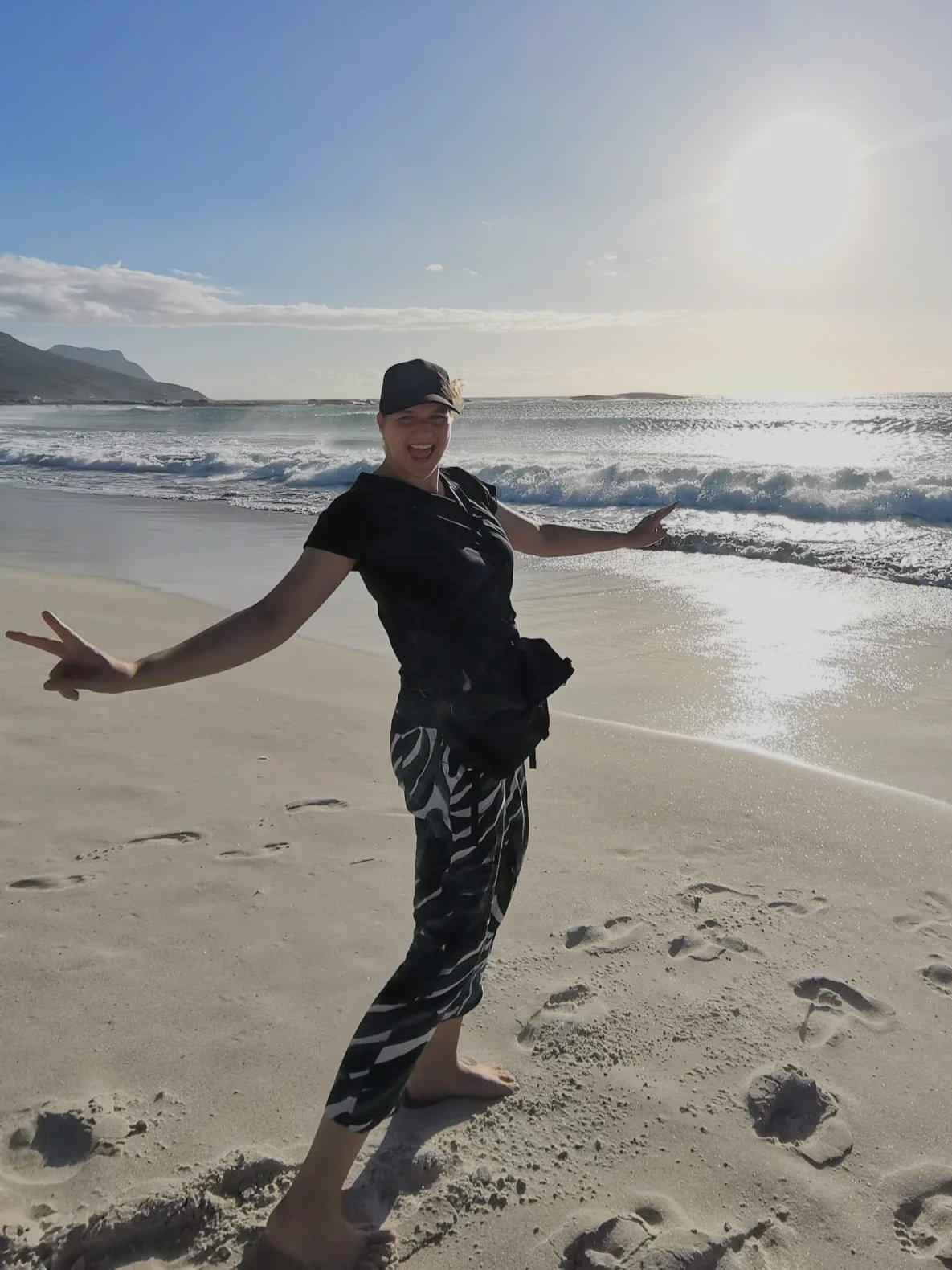 Eine lächelnde Frau steht barfuß am Strand in der Sonne, mit Blick auf das Meer, trägt schwarze Kleidung und macht Peace-Zeichen mit beiden Händen.