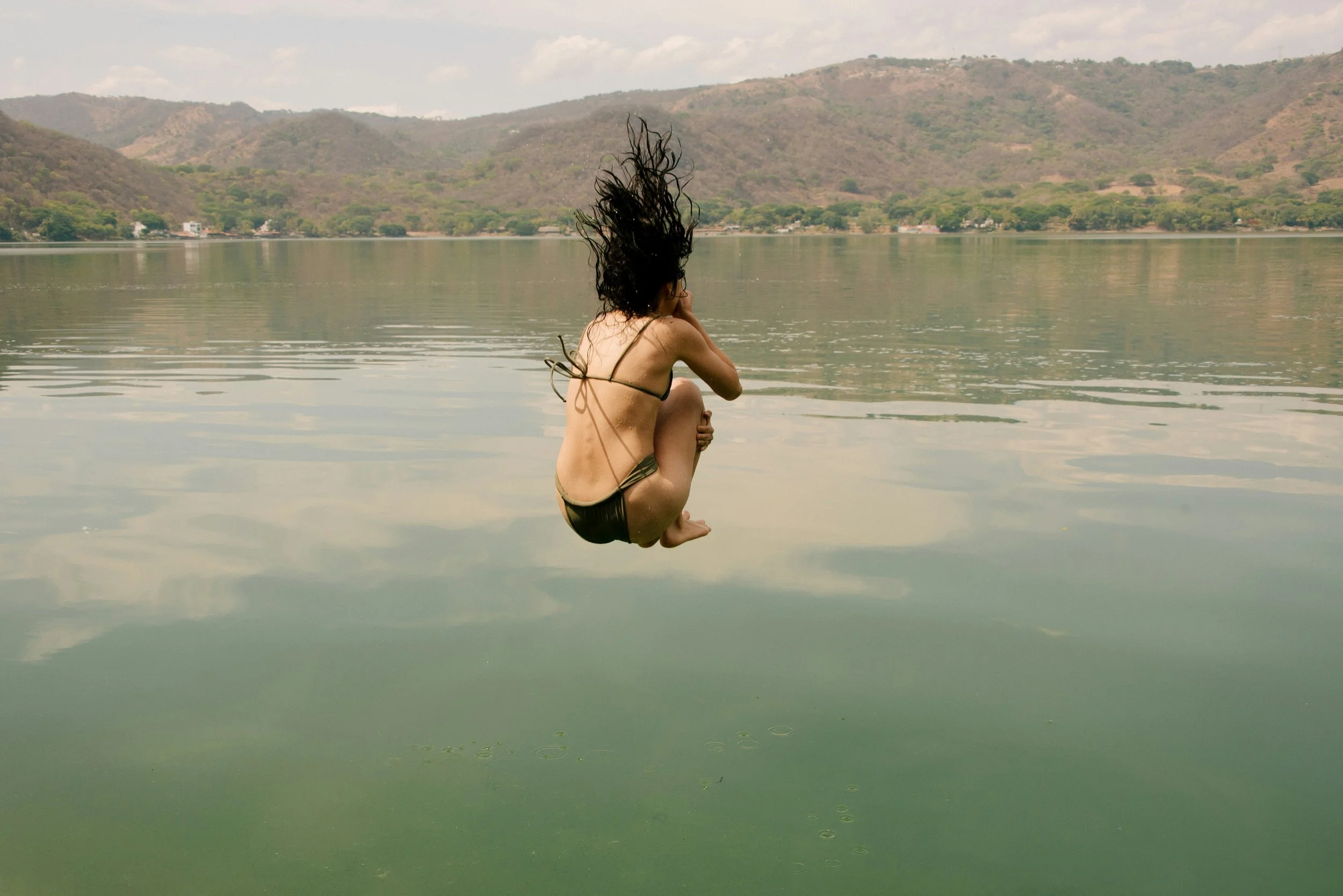 eine Frau in einem Bikino springt in den See, während Sie sich mit einer Hand die Nase zuhält