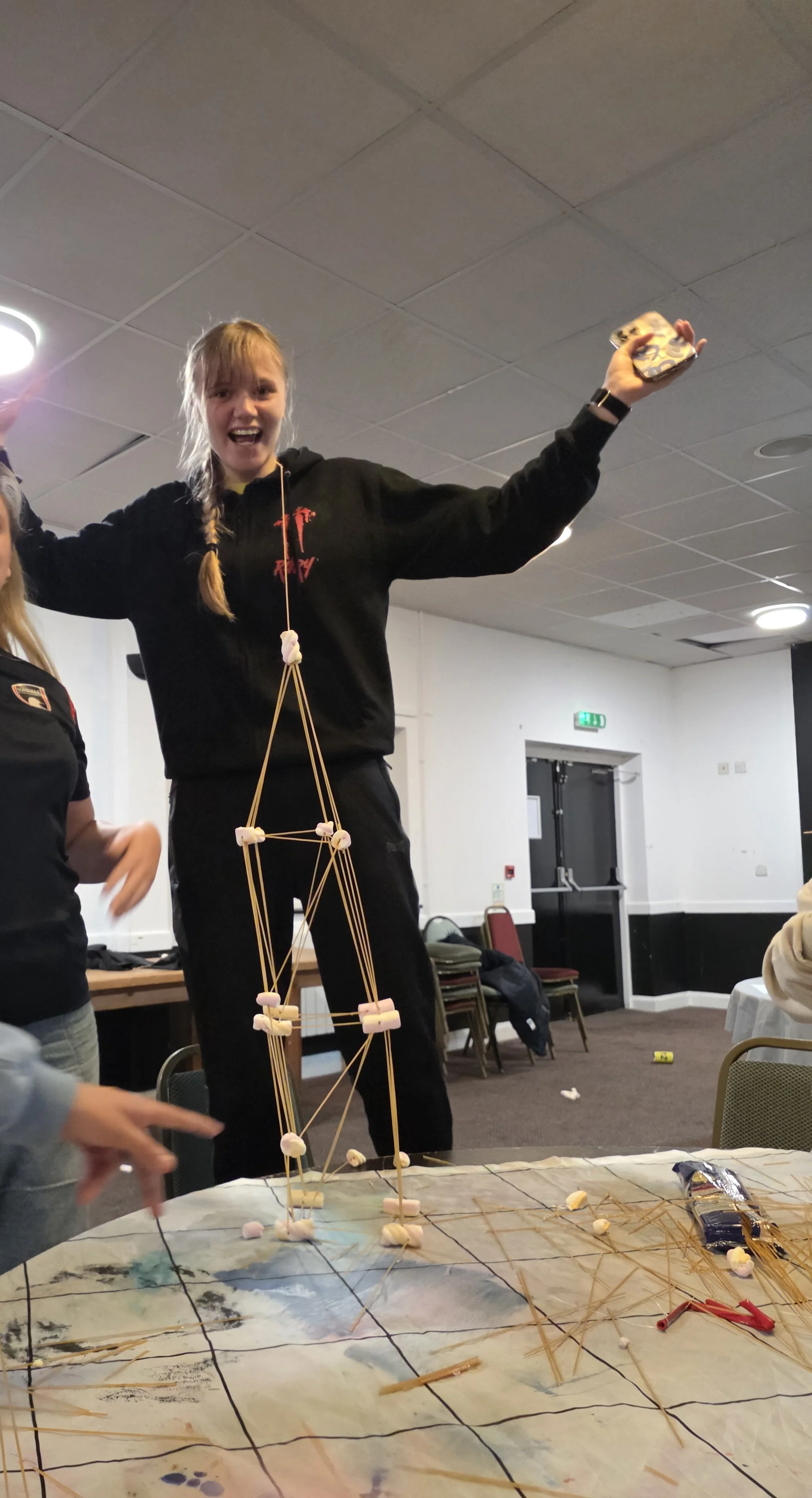 A young woman standing on a table, smiling and raising her arms, with a structure made of marshmallows and spaghetti noodles in front of her. She appears to be celebrating after completing a project, possibly a bridge or tower for a classroom activity.