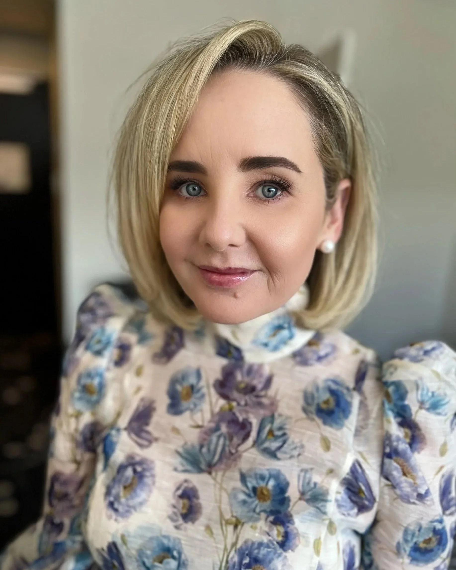 Close-up of a woman with blonde hair, blue eyes, and pearl earrings, wearing a floral-patterned top or dress.