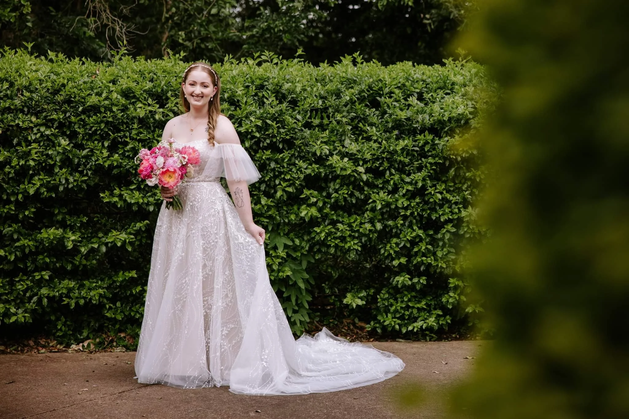 A smiling bride in a white lace wedding dress holding a pink bouquet, standing outdoors in front of a hedge.