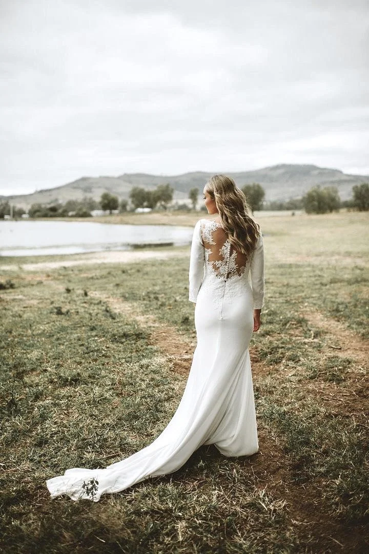 A bride standing outdoors on a grassy field near a body of water, wearing a long white wedding dress with lace details on the back, looking away from the camera with mountains and trees in the background.