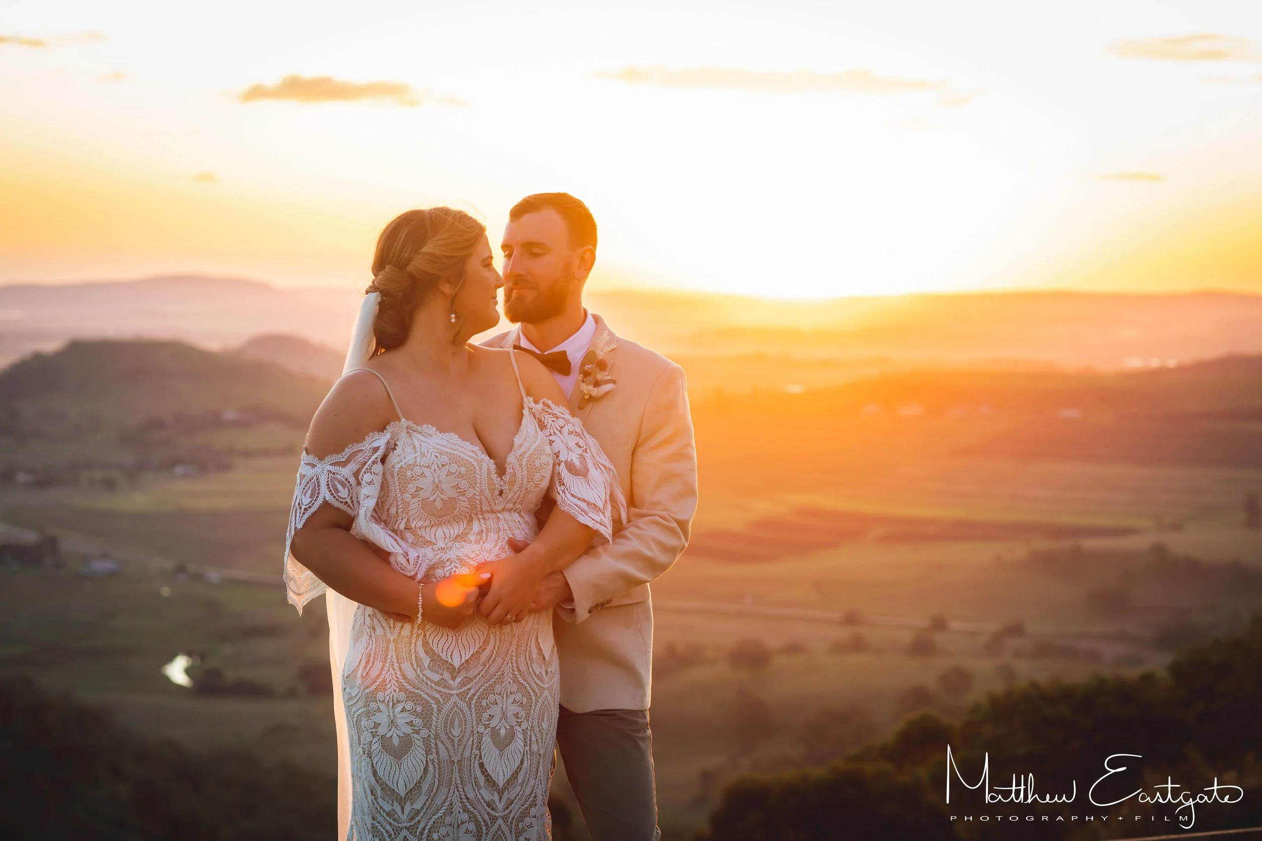 A bride and groom embrace each other during sunset outdoors, with rolling hills and a scenic sky in the background.
