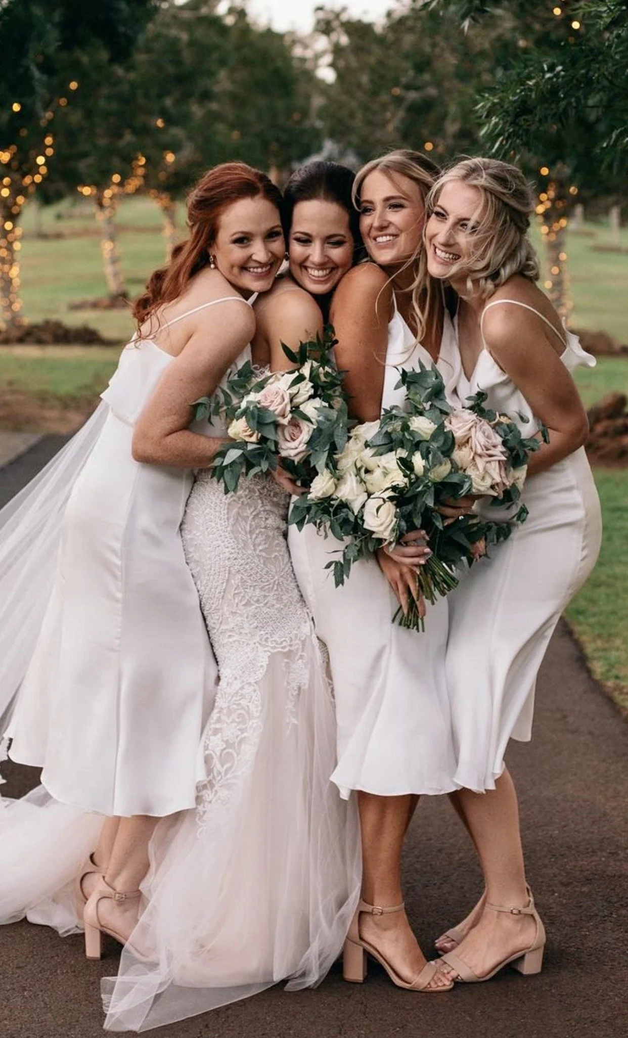 Four women in white dresses standing close together, smiling and holding bouquets of pink and white roses with green leaves, outdoors with trees and string lights in the background.