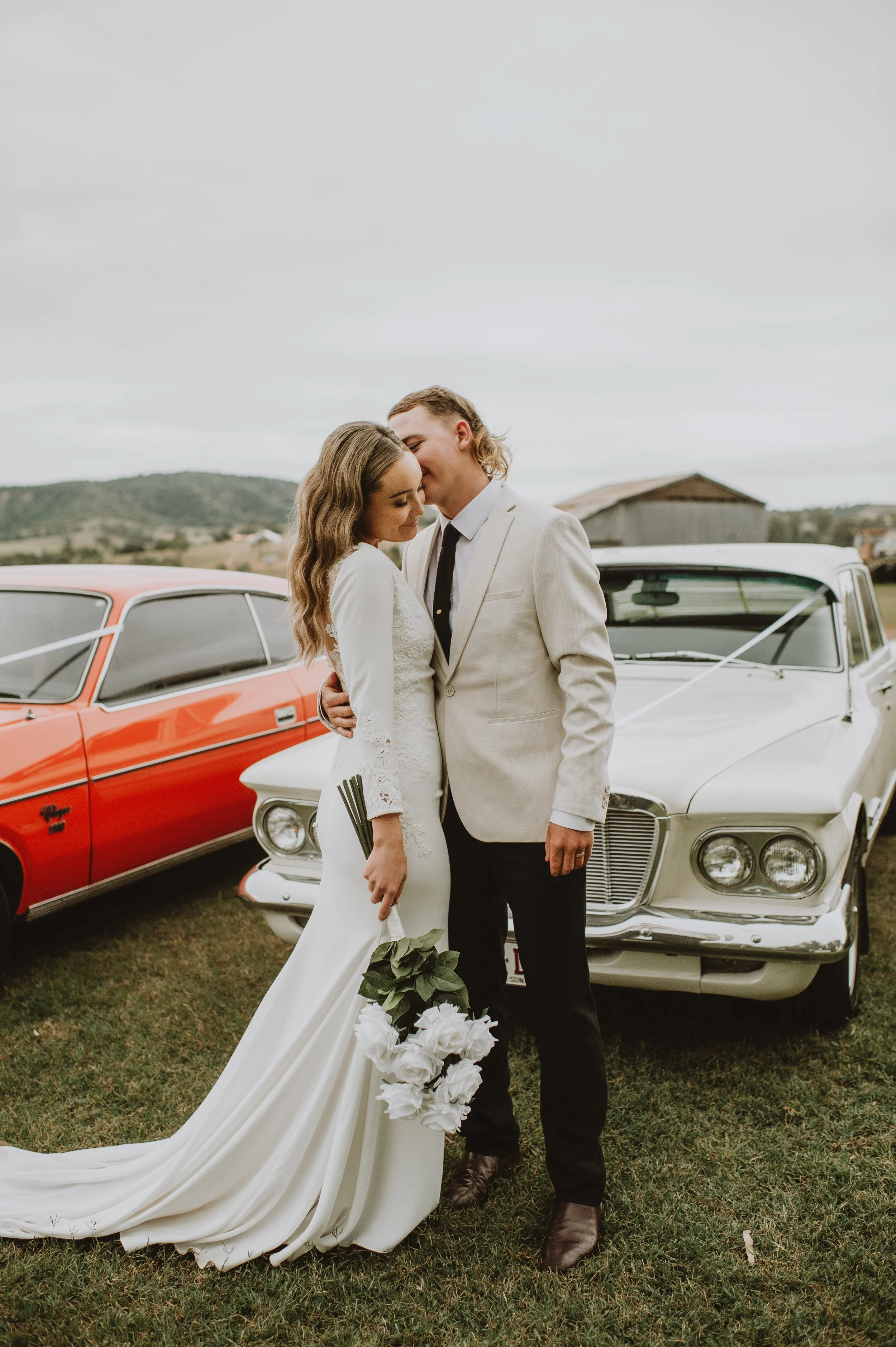 A bride and groom standing close together outdoors, with vintage cars and a rural landscape in the background, on a cloudy day.