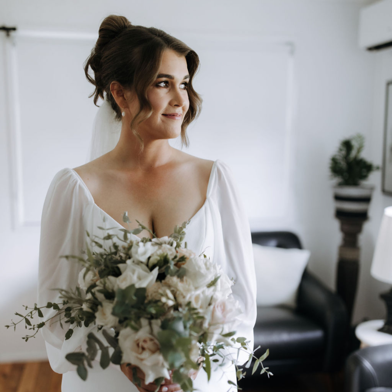 A bride holding a bouquet of white flowers and greenery, standing in a bright room, looking to her right with a gentle smile.