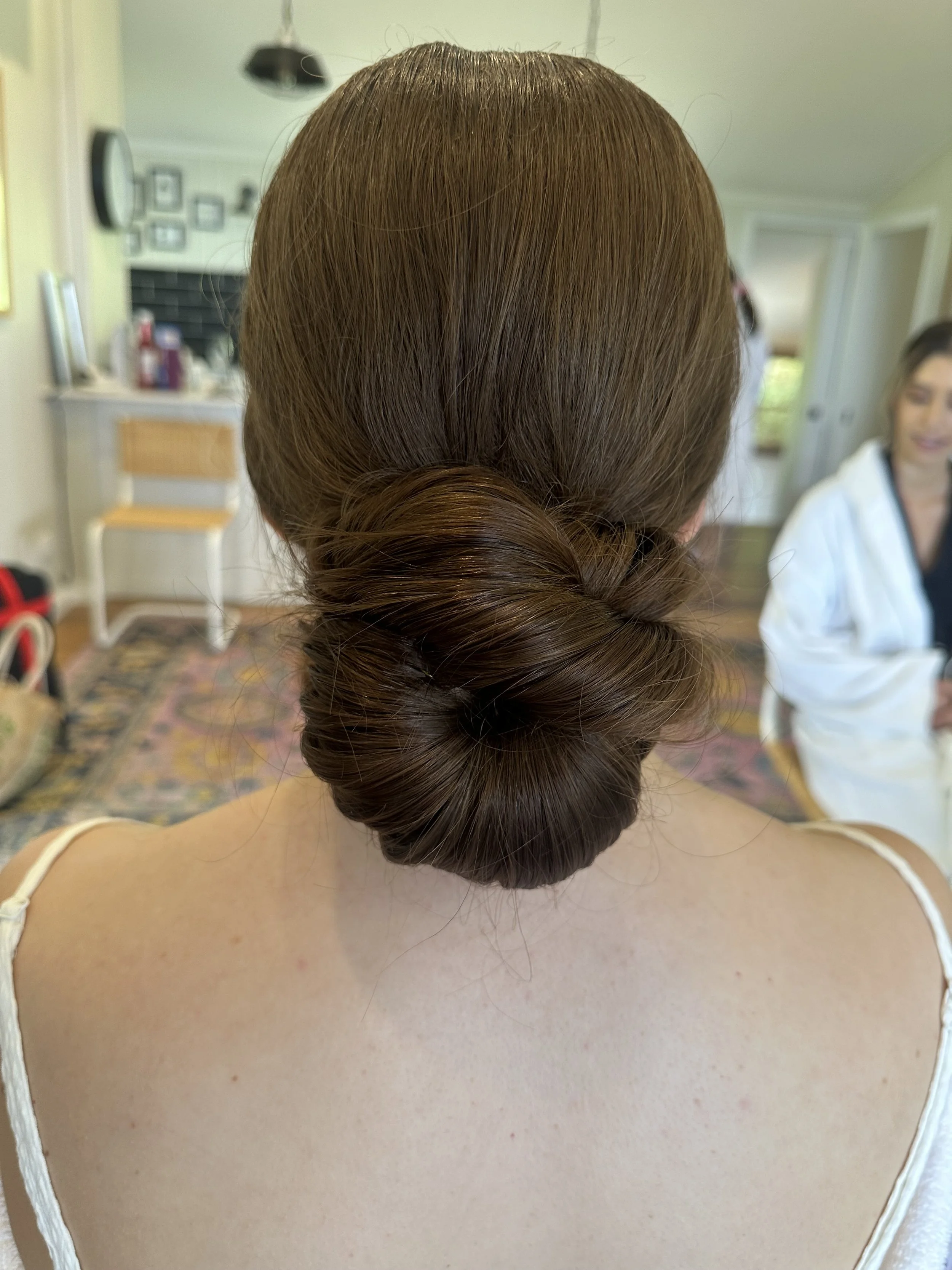 Back view of a woman with brown hair styled in a low, braided bun, wearing a white spaghetti strap top, in an indoor setting.