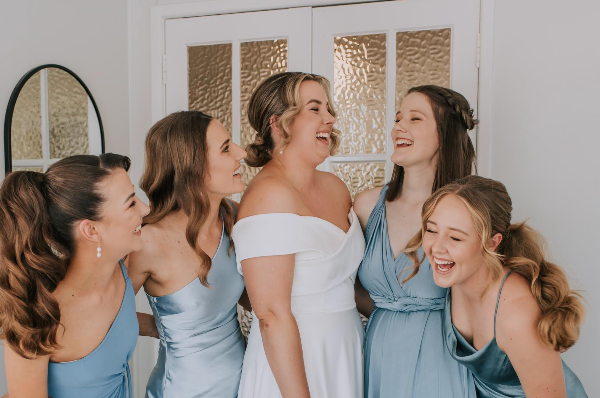 A bride in a white dress smiles as she is surrounded by bridesmaids in blue dresses, all appearing happy and joyful.