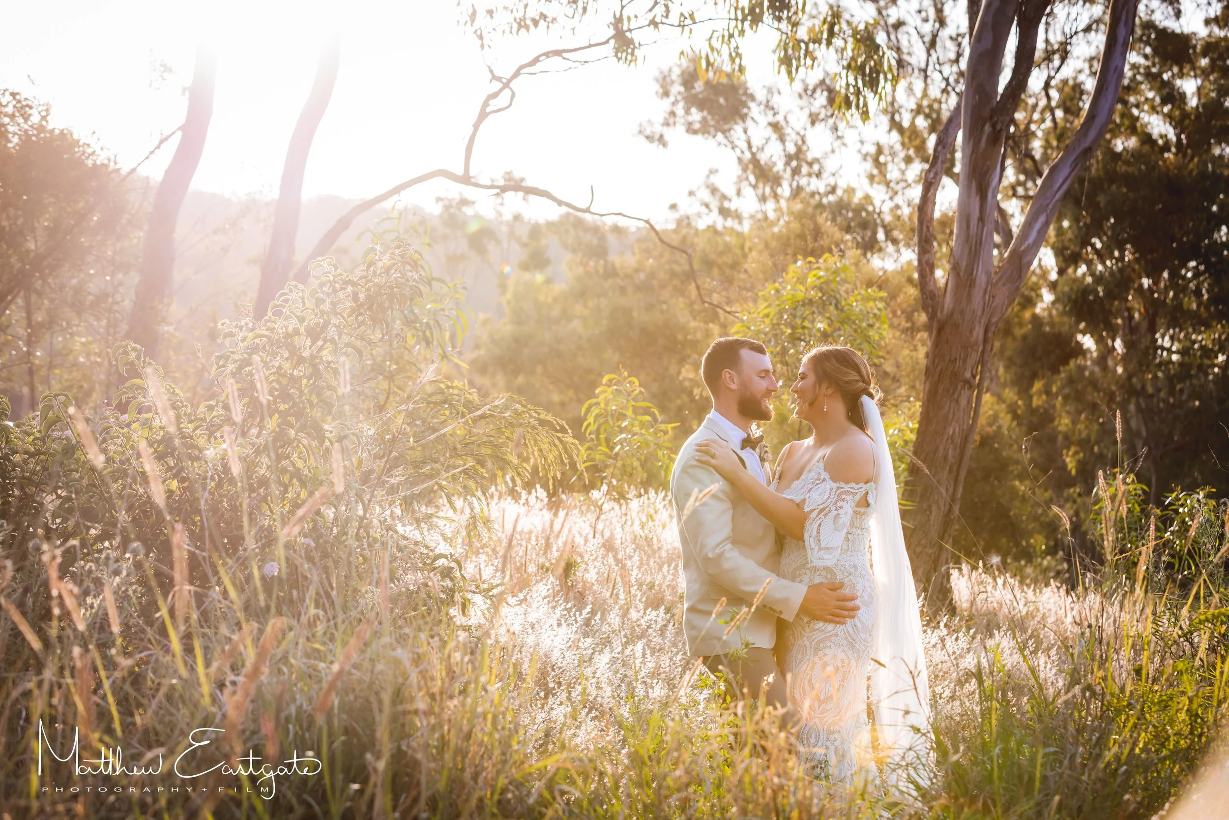 A couple dressed in wedding attire embracing outdoors near a lake, with trees and sunlight in the background.