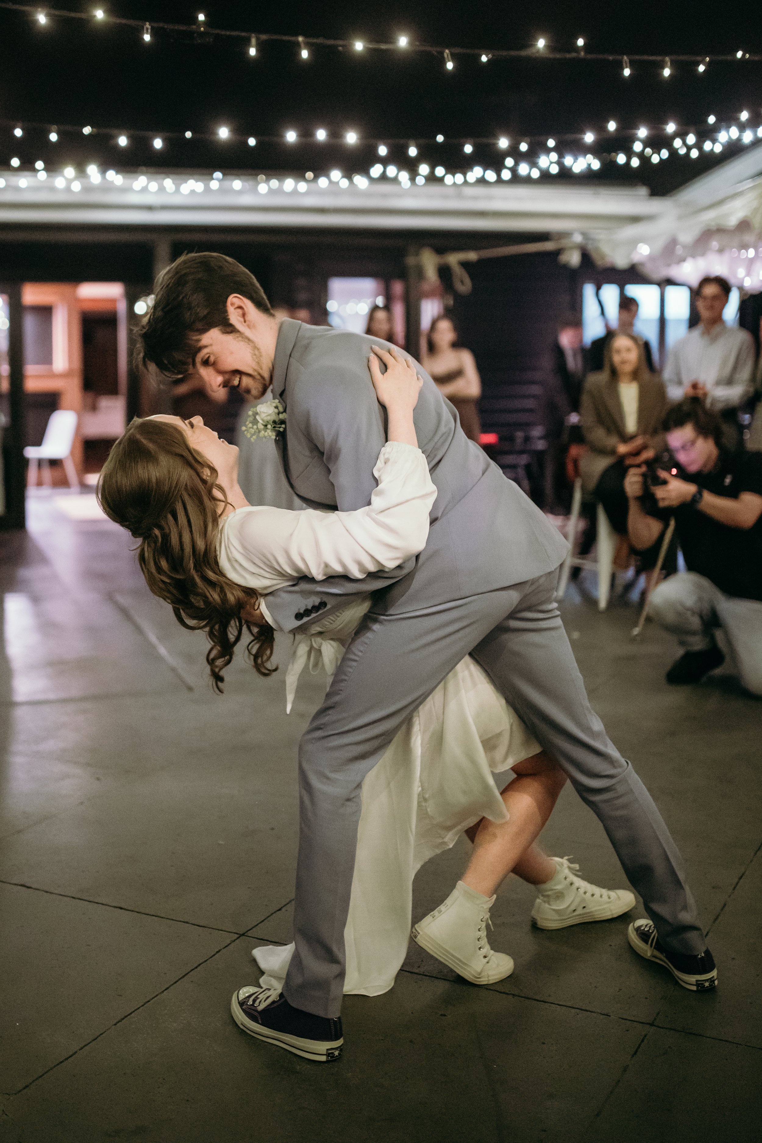 A couple dancing closely at a party, with a man in a gray suit and a woman in a white dress, surrounded by onlookers and string lights.