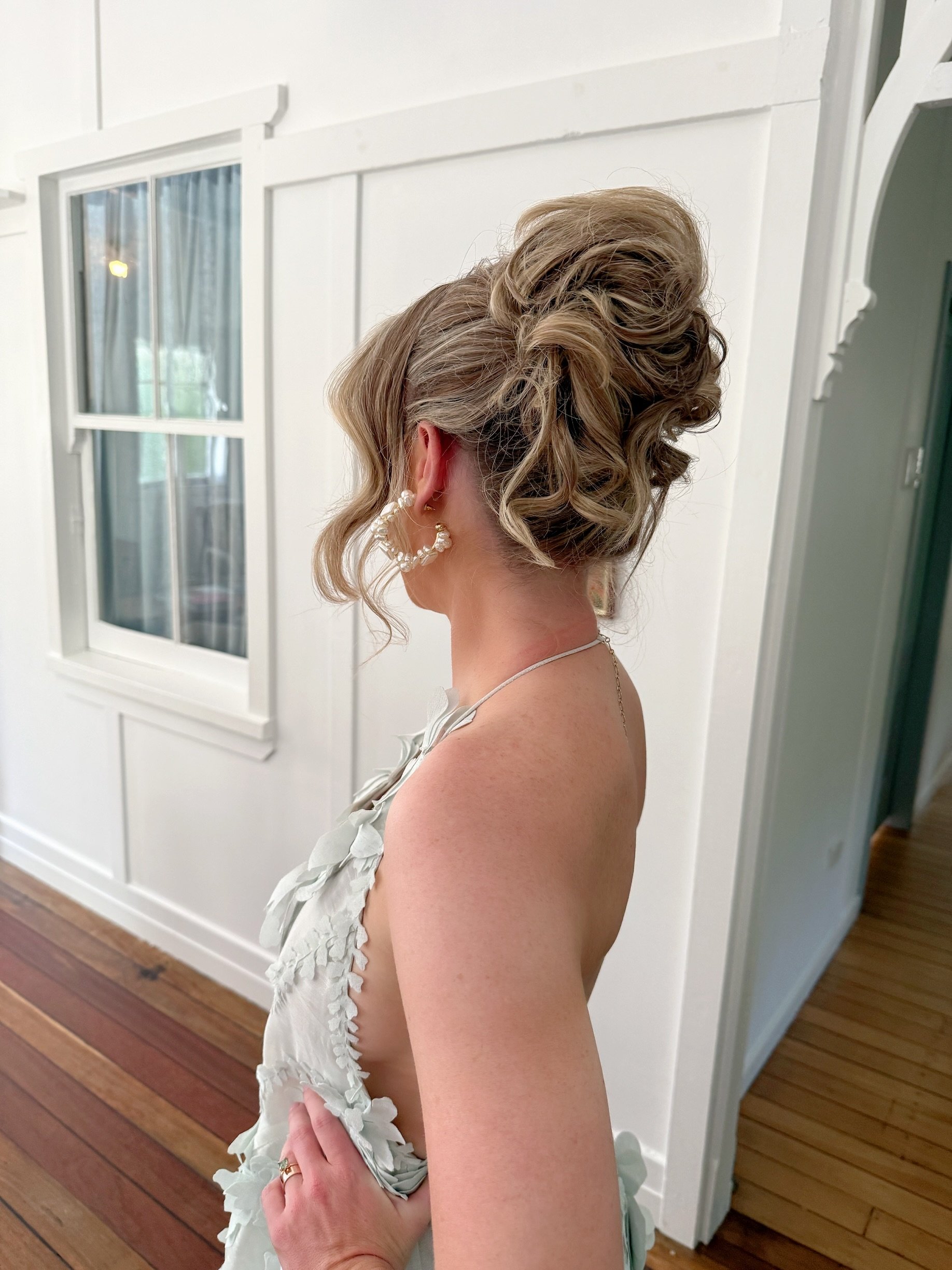 A woman with curly updo hairstyle wearing pearl earrings and ivory dress, standing indoors near a white wall and window.