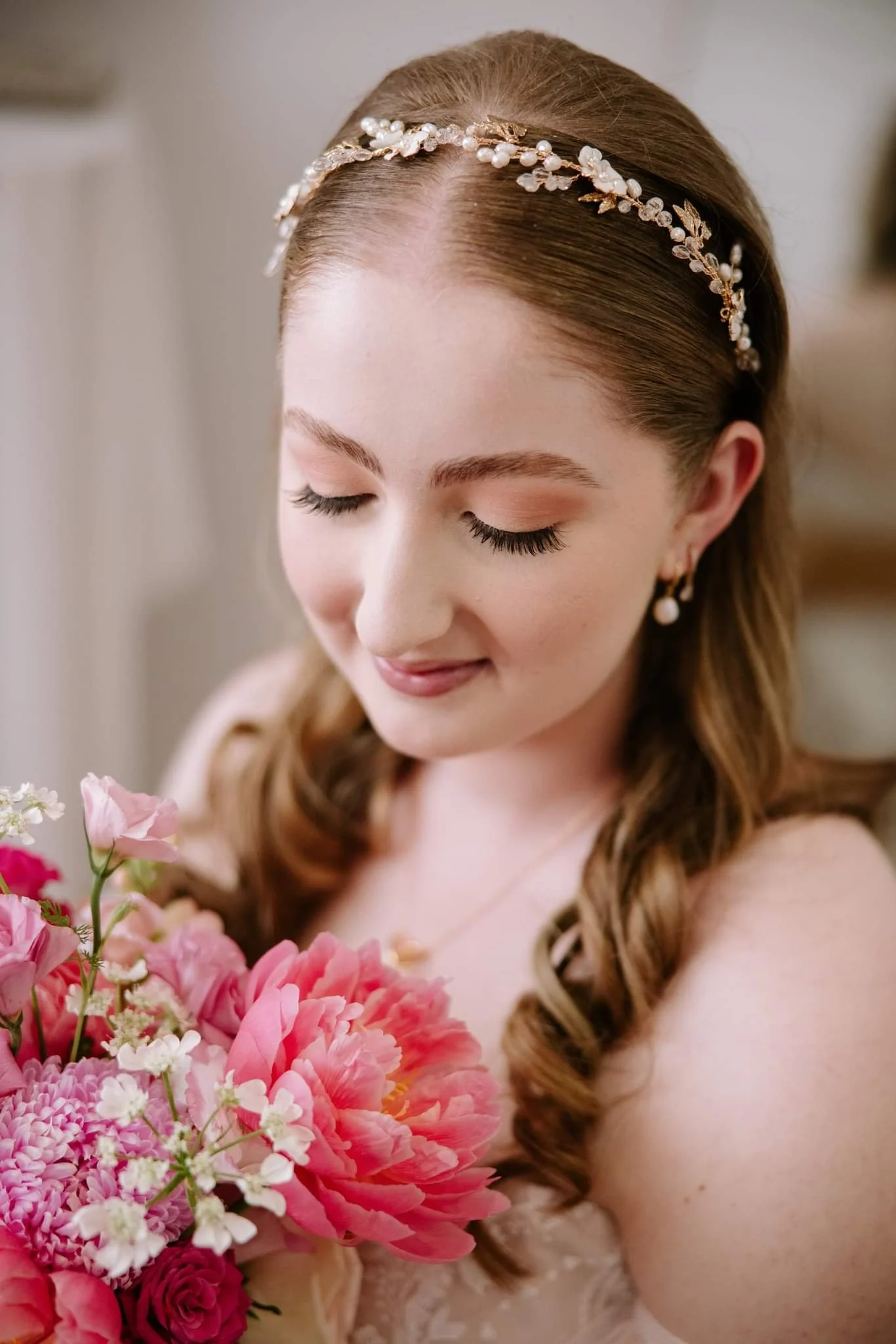 Close-up of a woman with auburn hair styled in loose curls, wearing a pearl and floral headband, holding a bouquet of pink and white flowers, with her eyes closed and a gentle expression.