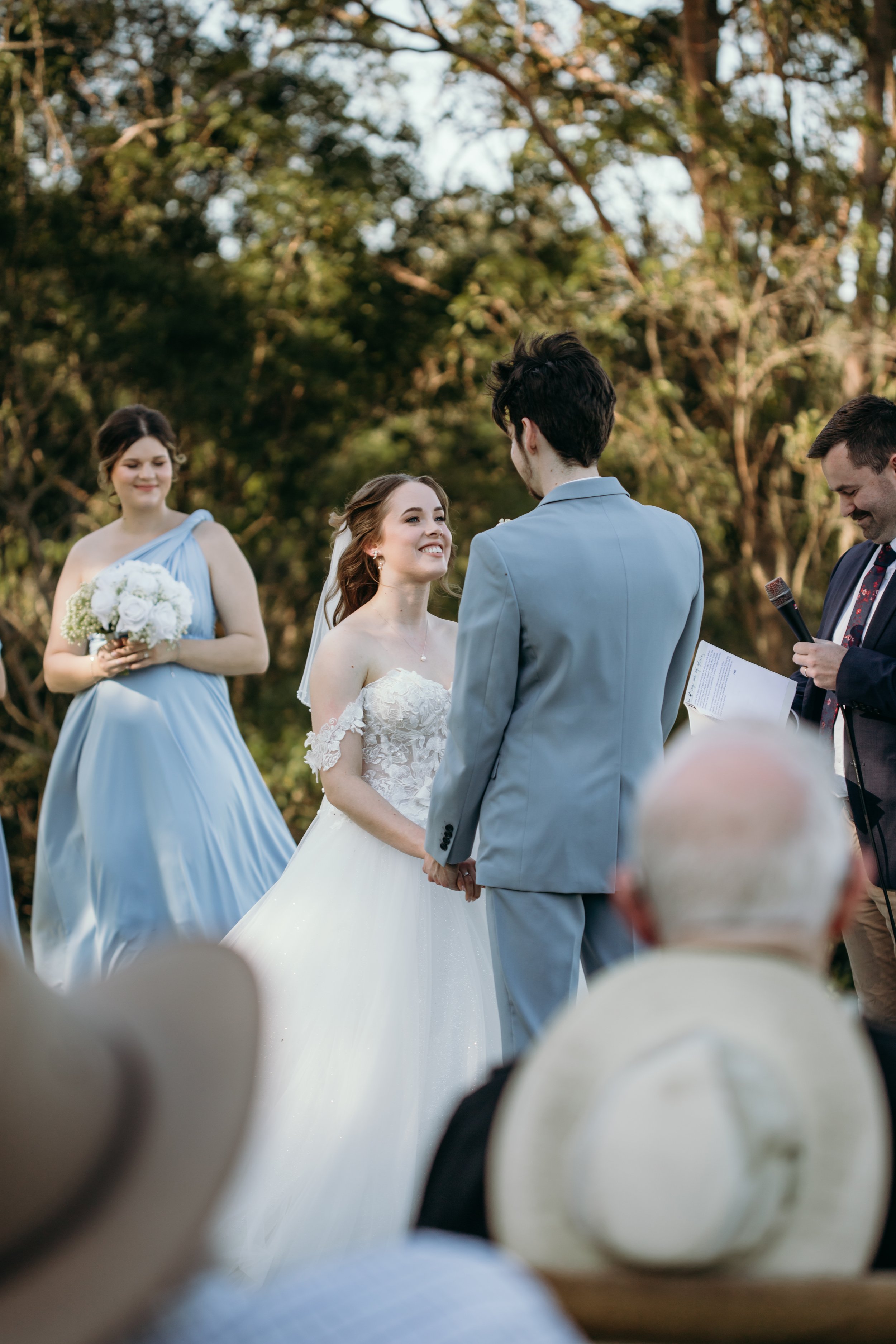 Bride and groom holding hands during outdoor wedding ceremony with officiant, bridesmaid holding bouquet, and guests in background.