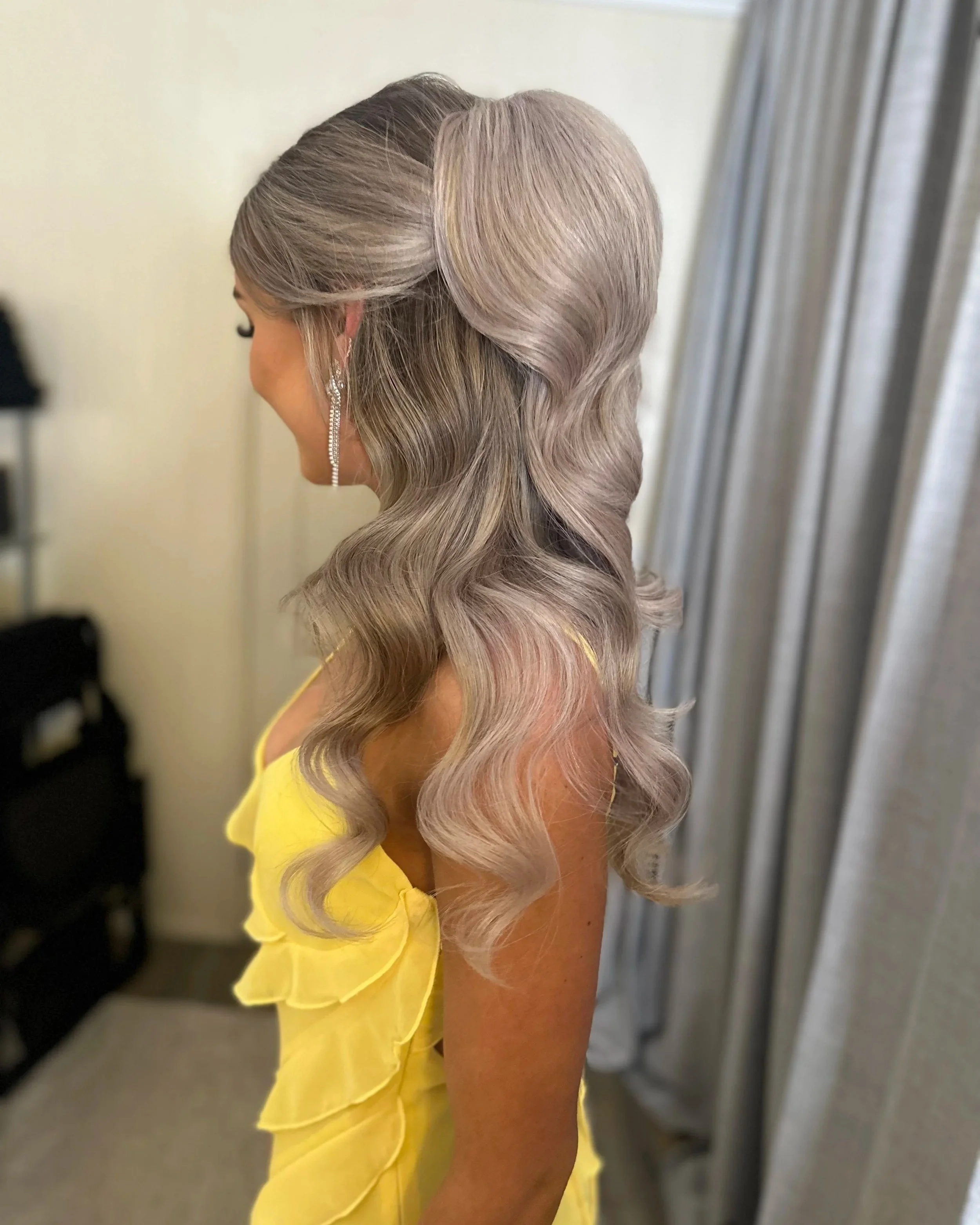 Side view of a woman with long, wavy, silver-gray hair wearing a yellow dress and silver drop earrings, standing in front of a background with vertical blinds.