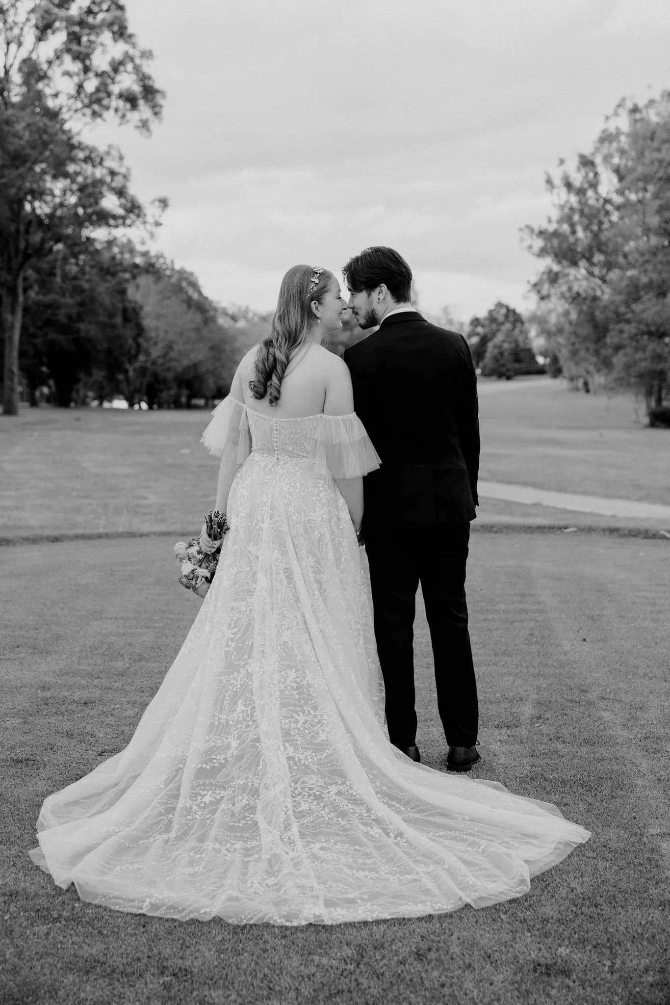 A black and white photo of a bride and groom standing in a park, face to face with noses touching, trees in the background, the bride in a lace wedding gown holding a bouquet, and the groom in a suit.