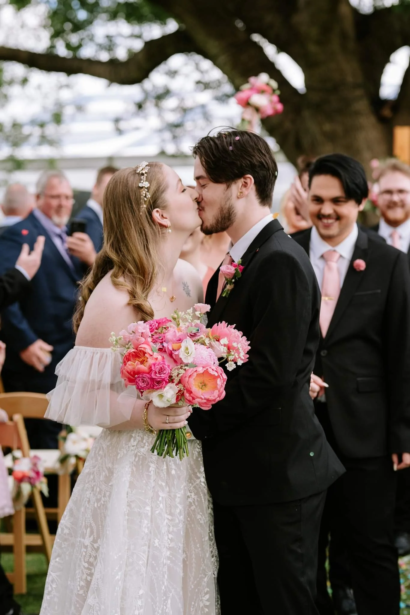 A bride and groom are kissing during their wedding ceremony surrounded by friends and family, with the bride holding a bouquet of pink and white flowers.