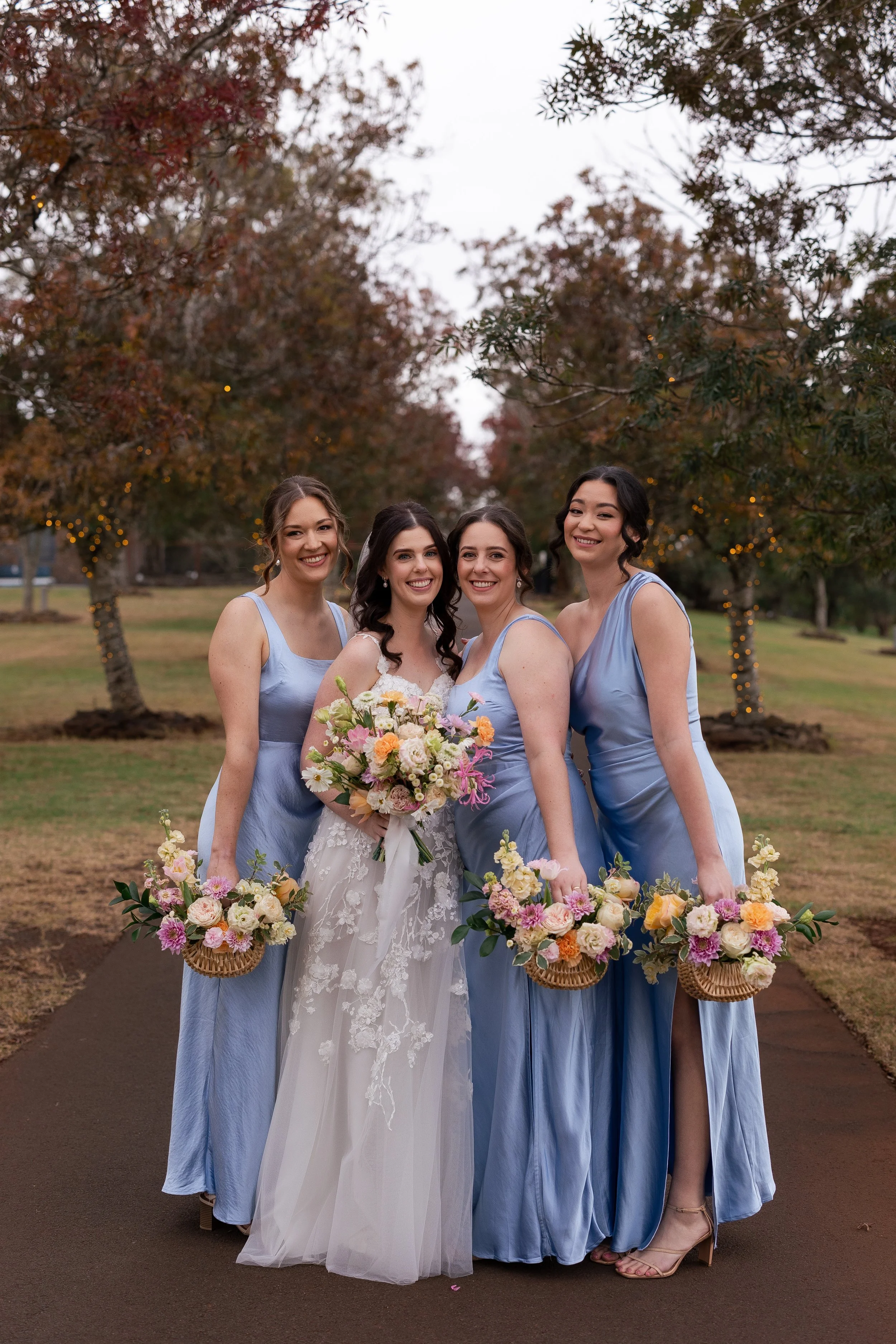 Group of five women, one in a white wedding dress and the others in blue dresses, standing outdoors on a paved pathway during early evening. They are holding bouquets of flowers, and trees with autumn leaves and string lights are visible in the backg