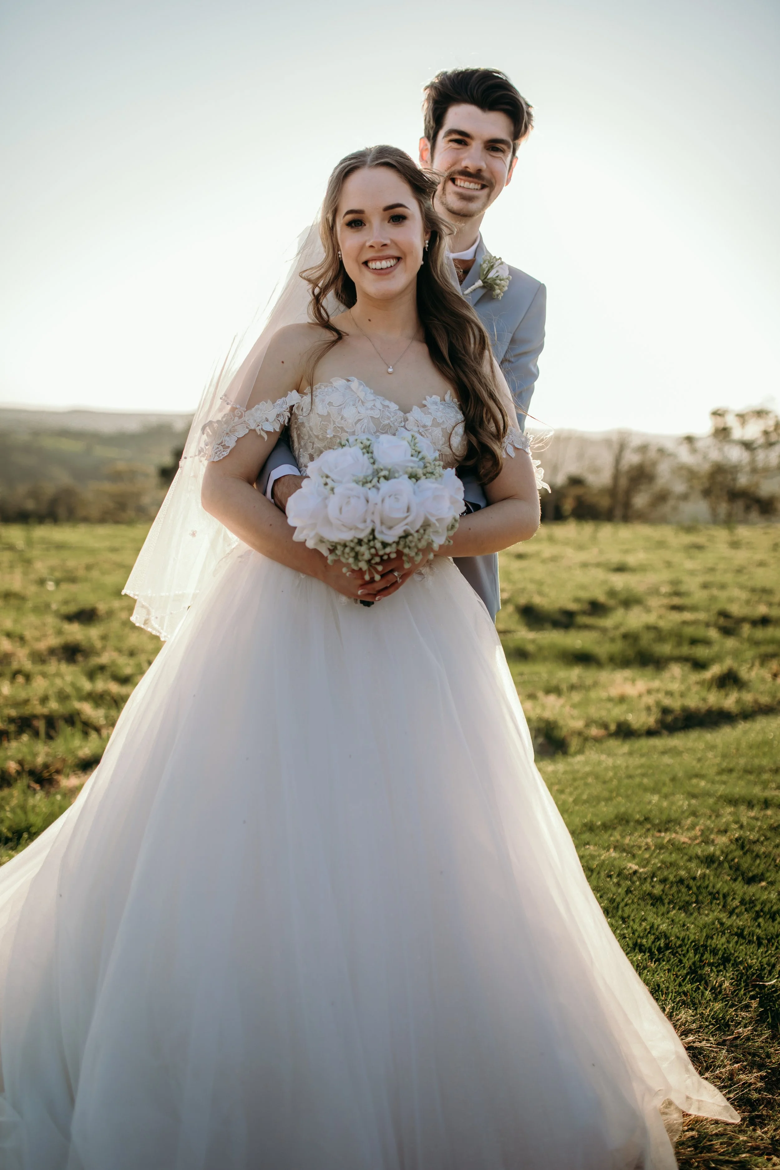 Bride and groom smiling outdoors, bride holding a bouquet of white roses, wedding dress with off-shoulder lace details, groom in a light gray suit with a boutonniere, open field background with trees and hills under a clear sky.