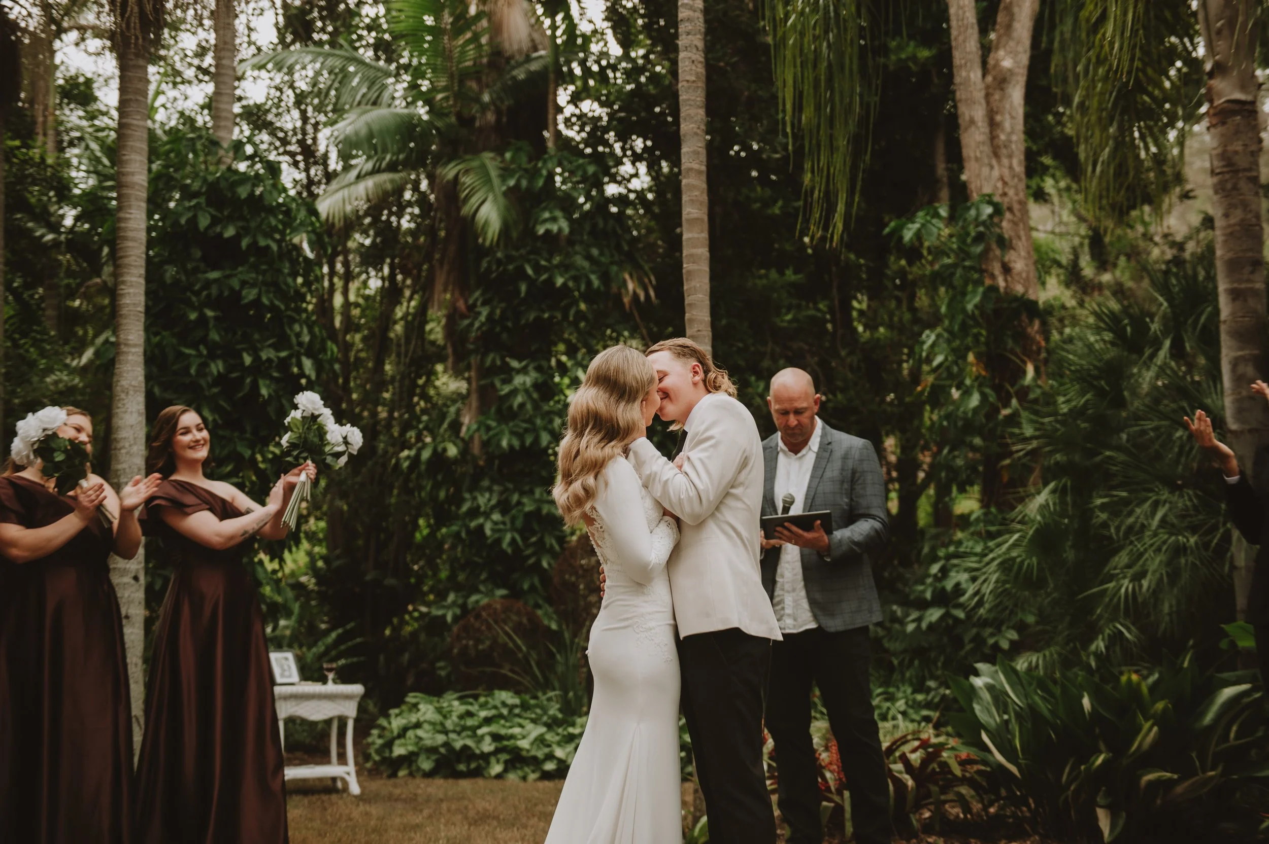 A couple sharing a kiss during their outdoor wedding ceremony with friends and family in a lush, tropical garden.