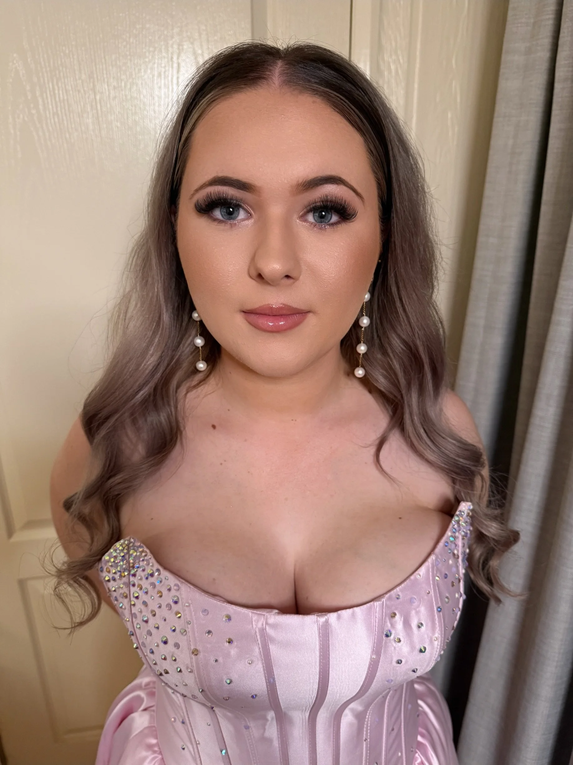 A woman with long, wavy brown hair, wearing a pink dress with sparkling embellishments and pearl earrings, standing indoors against a beige wall and curtain.