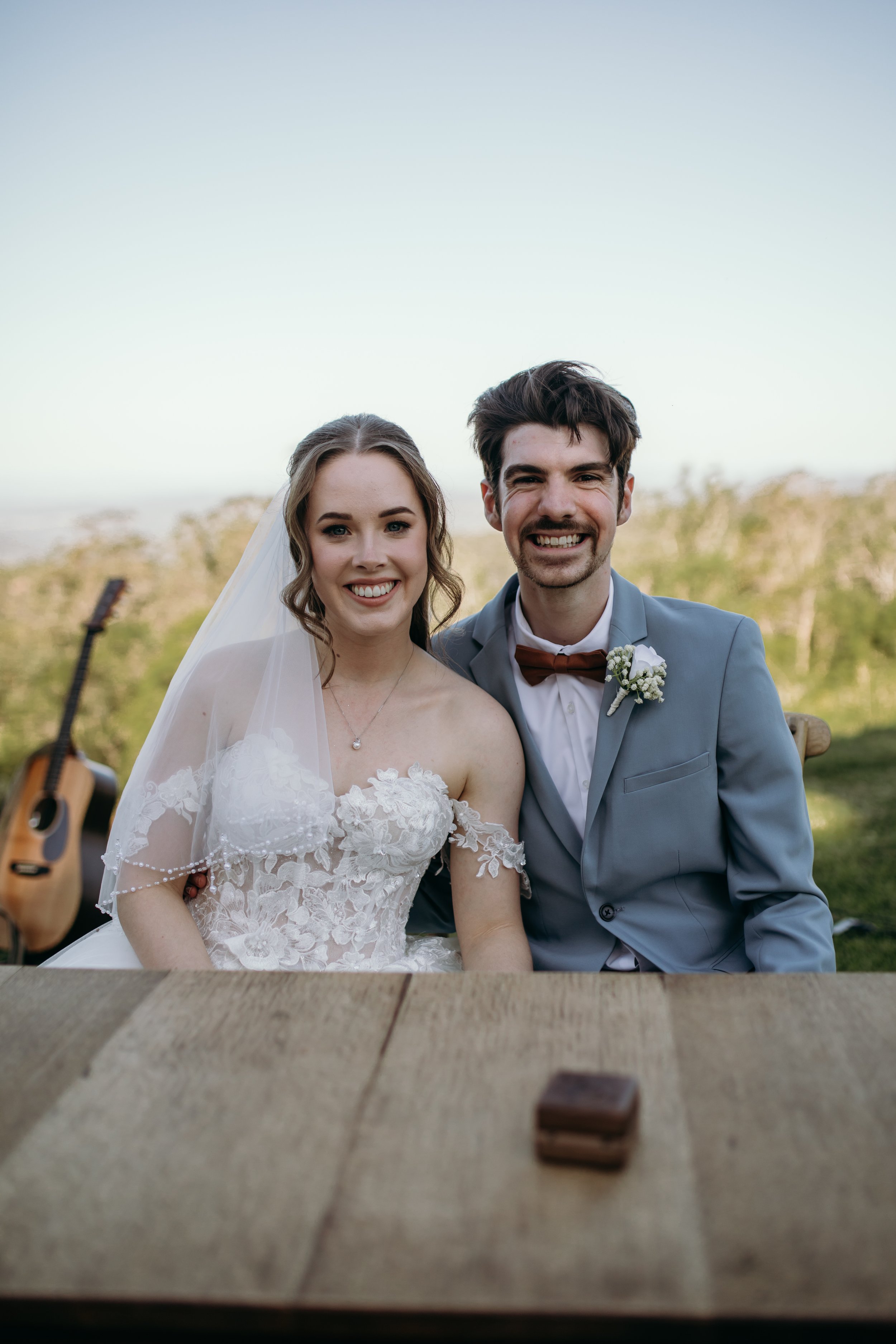 A smiling bride and groom sitting at a wooden table outdoors, with a guitar and a small box on the table. They are dressed in wedding attire, with the bride in a white lace gown and veil, and the groom in a gray suit and maroon bow tie, with greenery