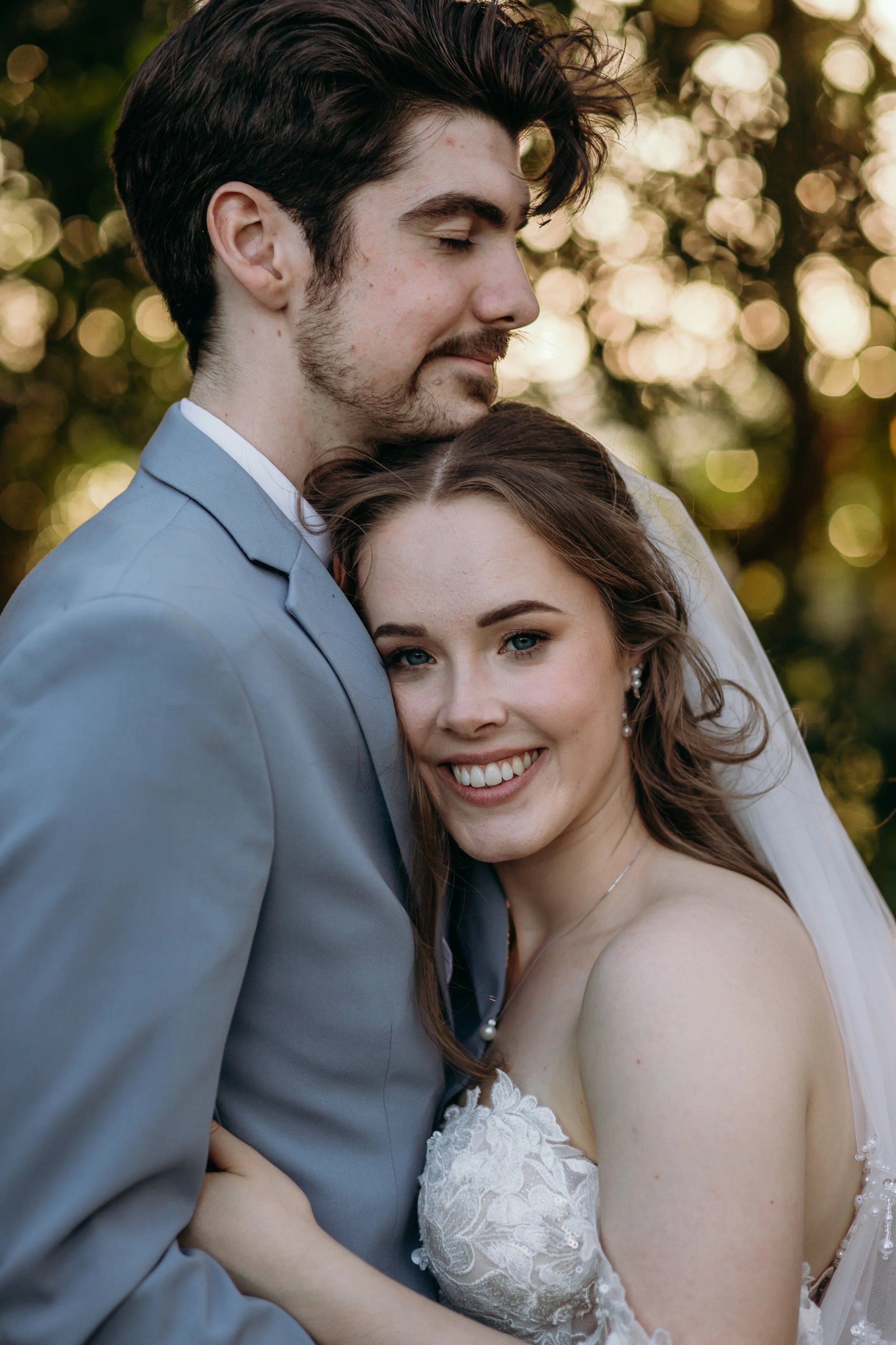 A close-up of a newlywed couple hugging outdoors, with the groom in a light gray suit and the bride in a lace wedding dress smiling.