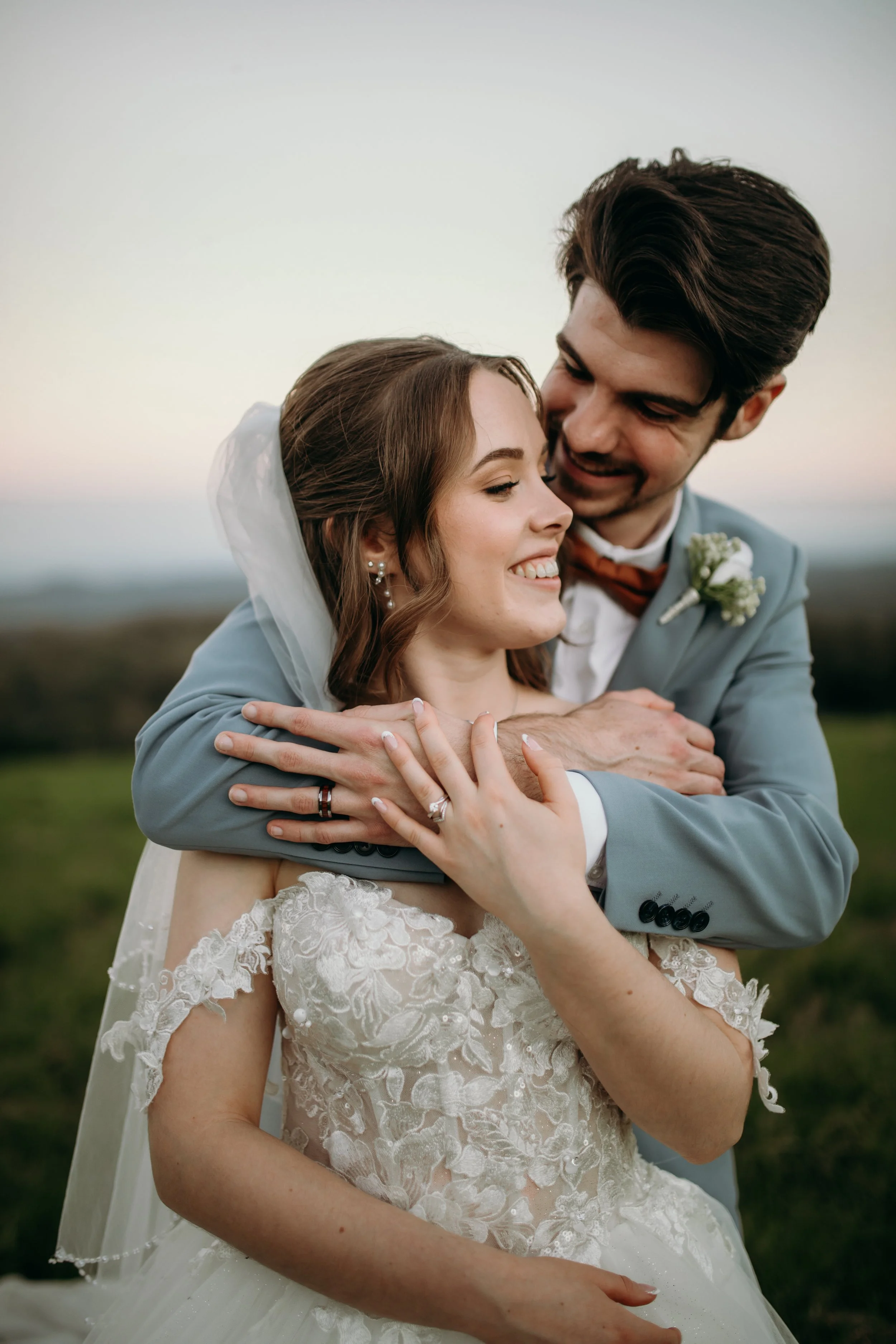 A bride and groom embracing outside during sunset. The bride wears a white lace wedding dress and veil, and the groom is in a light gray tuxedo with a white shirt and orange bow tie. They are smiling and looking at each other.