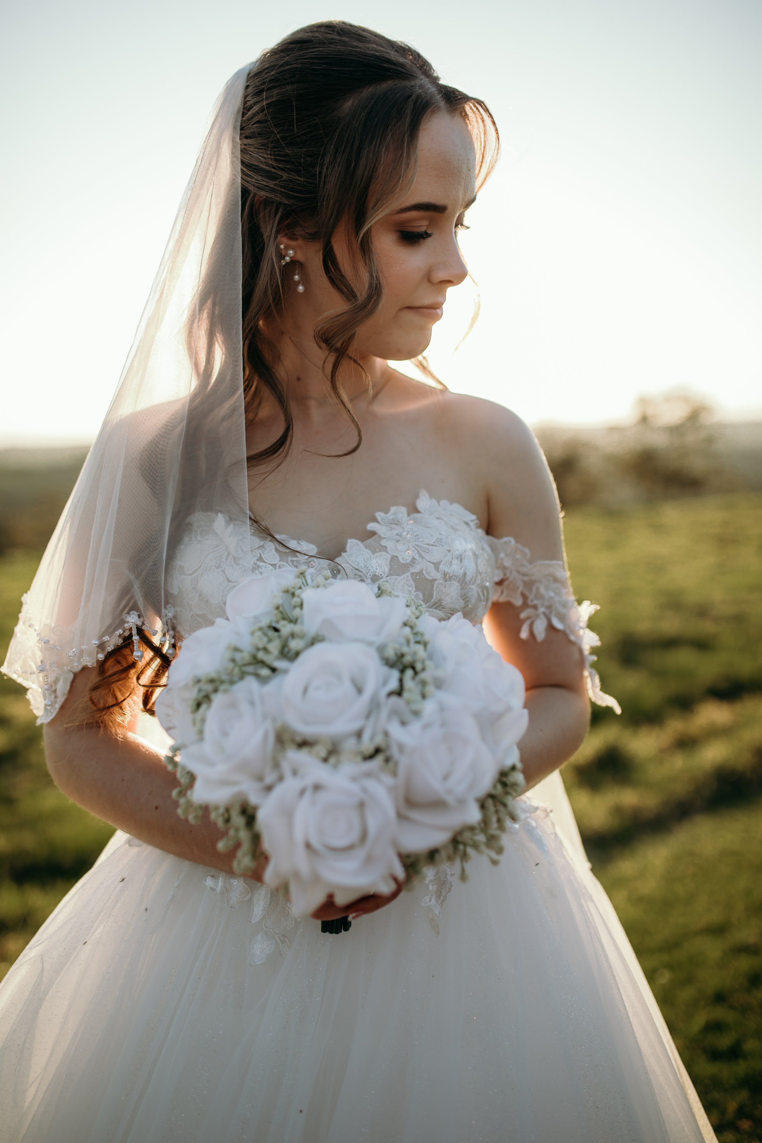 Bride holding a bouquet of white roses, wearing a lace wedding dress and veil, outdoors during sunset.