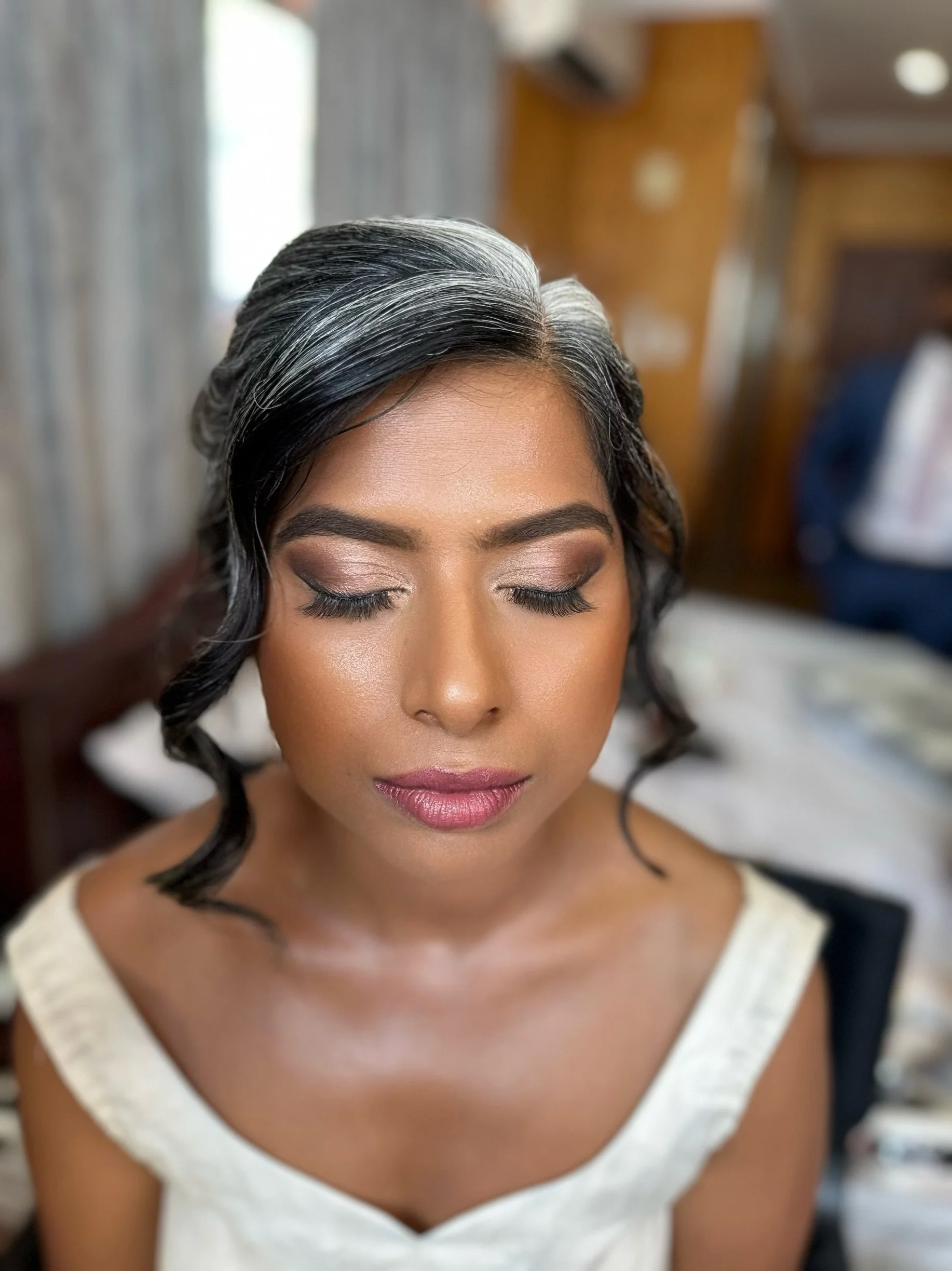 A woman with styled black and gray hair, closed eyes, and makeup, wearing a beige top with black straps, sitting indoors with blurred background.