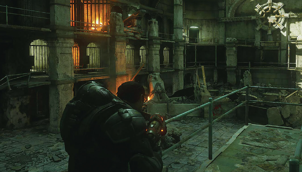 A person in futuristic armor holding a gun inside a damaged, industrial-style building with fires burning in the background and debris scattered around.