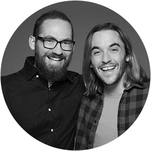 Two men smiling, one with a beard and glasses, the other with long hair, standing close together in front of a dark background.