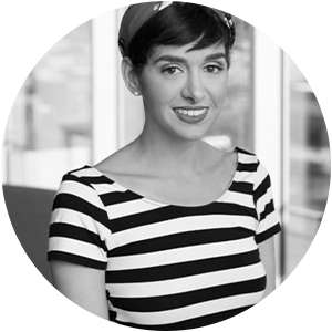 Black and white photo of a woman with short hair in a striped shirt, smiling indoors with windows in the background.