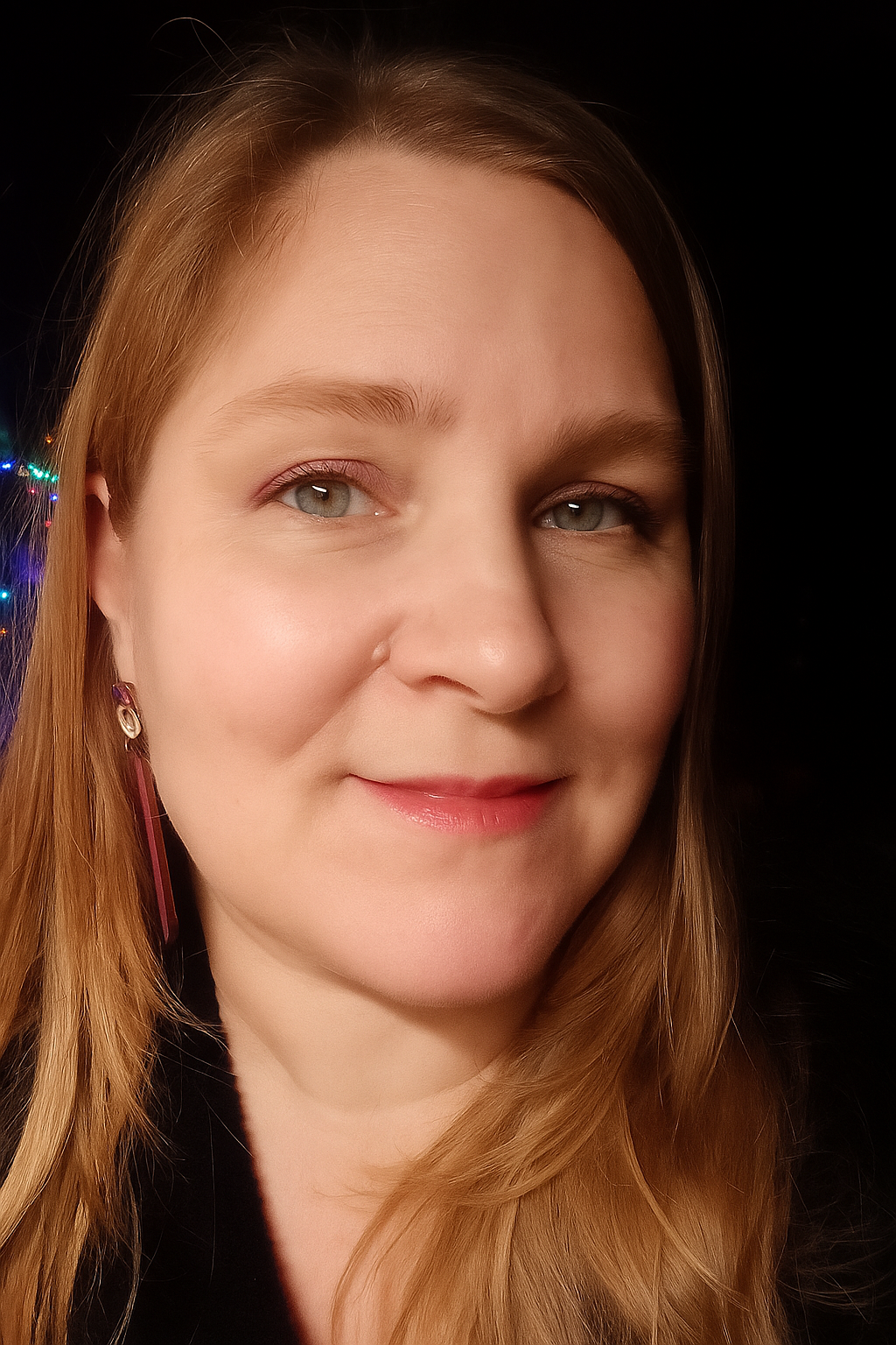 Close-up of a smiling woman with long dark blond hair and light-colored eyes, wearing earrings, against a dark background.