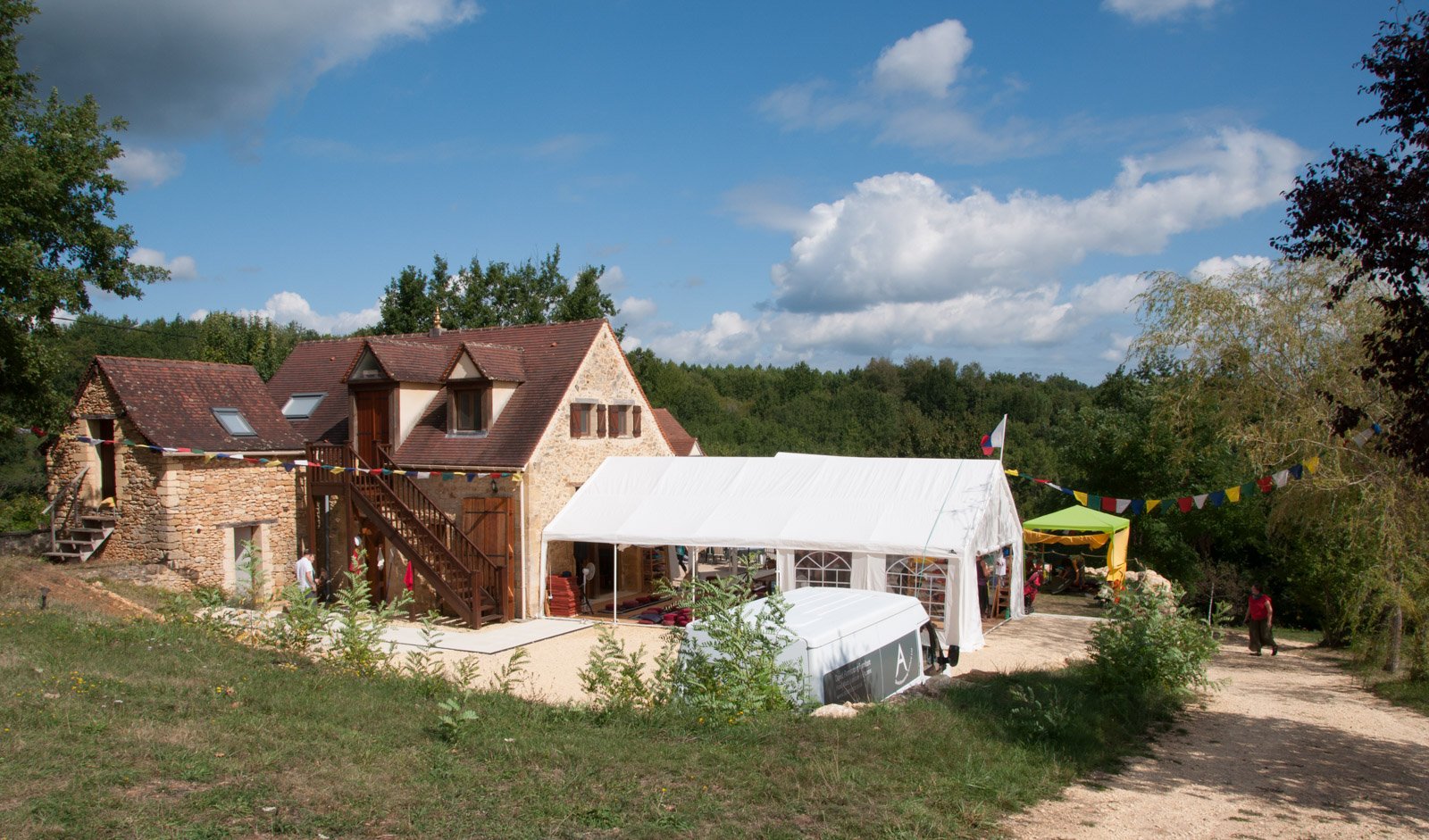 A rustic countryside house with a stone exterior and a red tile roof, surrounded by trees and a grassy area. A large white event tent is set up beside it, decorated with colorful pennant banners. There are a few people walking on a dirt path nearby, and a white van is parked in front of the tent.