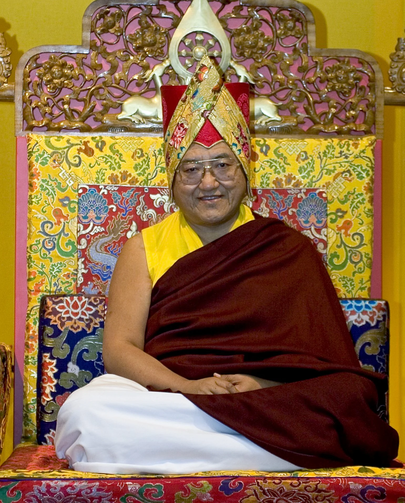 A Buddhist monk seated on an ornate, colorful throne with intricate floral designs, wearing a traditional multicolored turban, yellow shirt, maroon robe, and glasses, set against a bright yellow background.