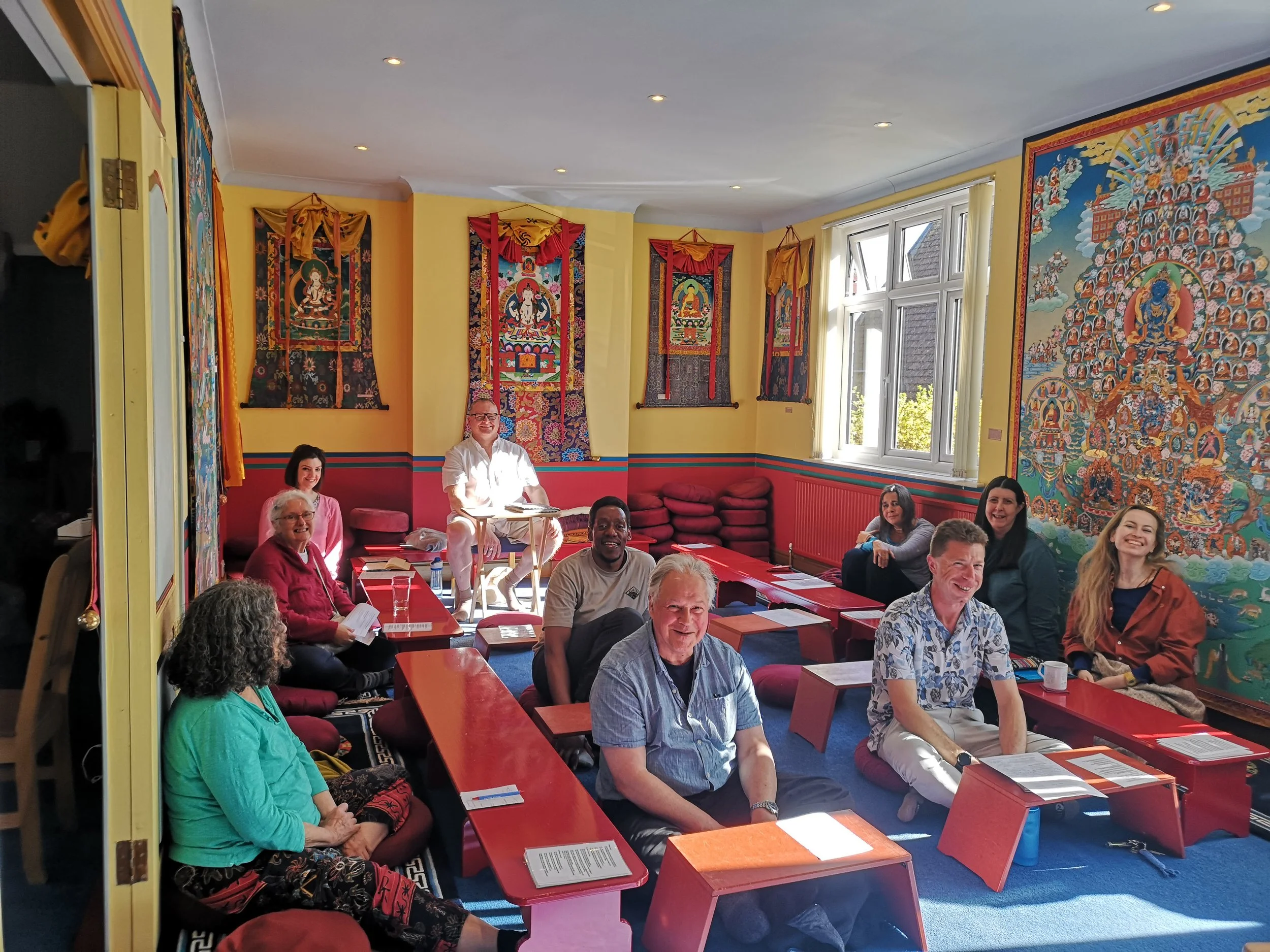 Group of people sitting on cushions and chairs in a brightly decorated room with yellow walls, featuring traditional Tibetan Thangka paintings and a large colorful tapestry on the wall.