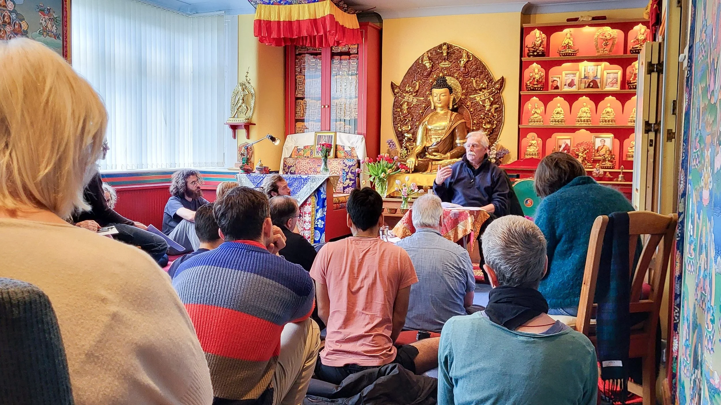 Group of people sitting on cushions and low tables in a room with colorful tapestries and Buddhist artwork on the walls, sunlight coming through the window.
