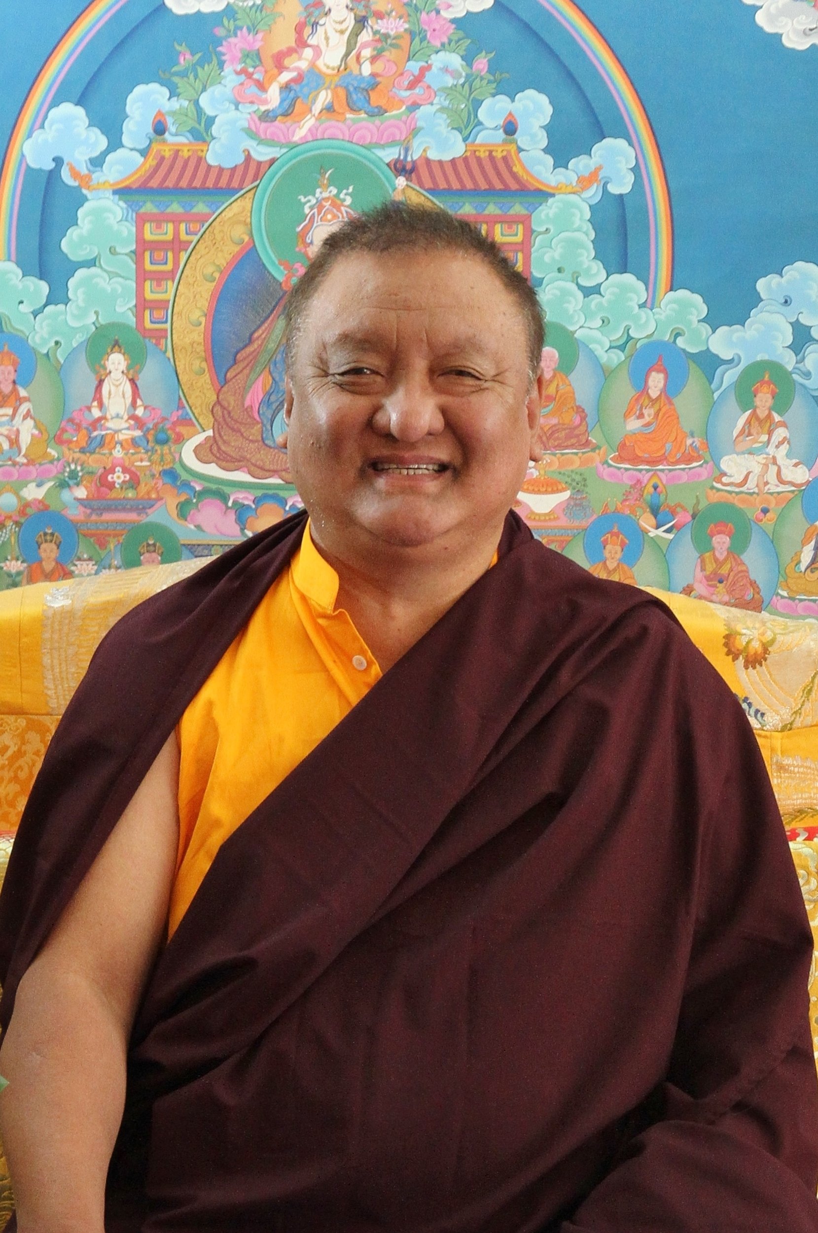 A Buddhist monk sitting in front of a colorful religious mural with deities and clouds, smiling and wearing traditional robes.
