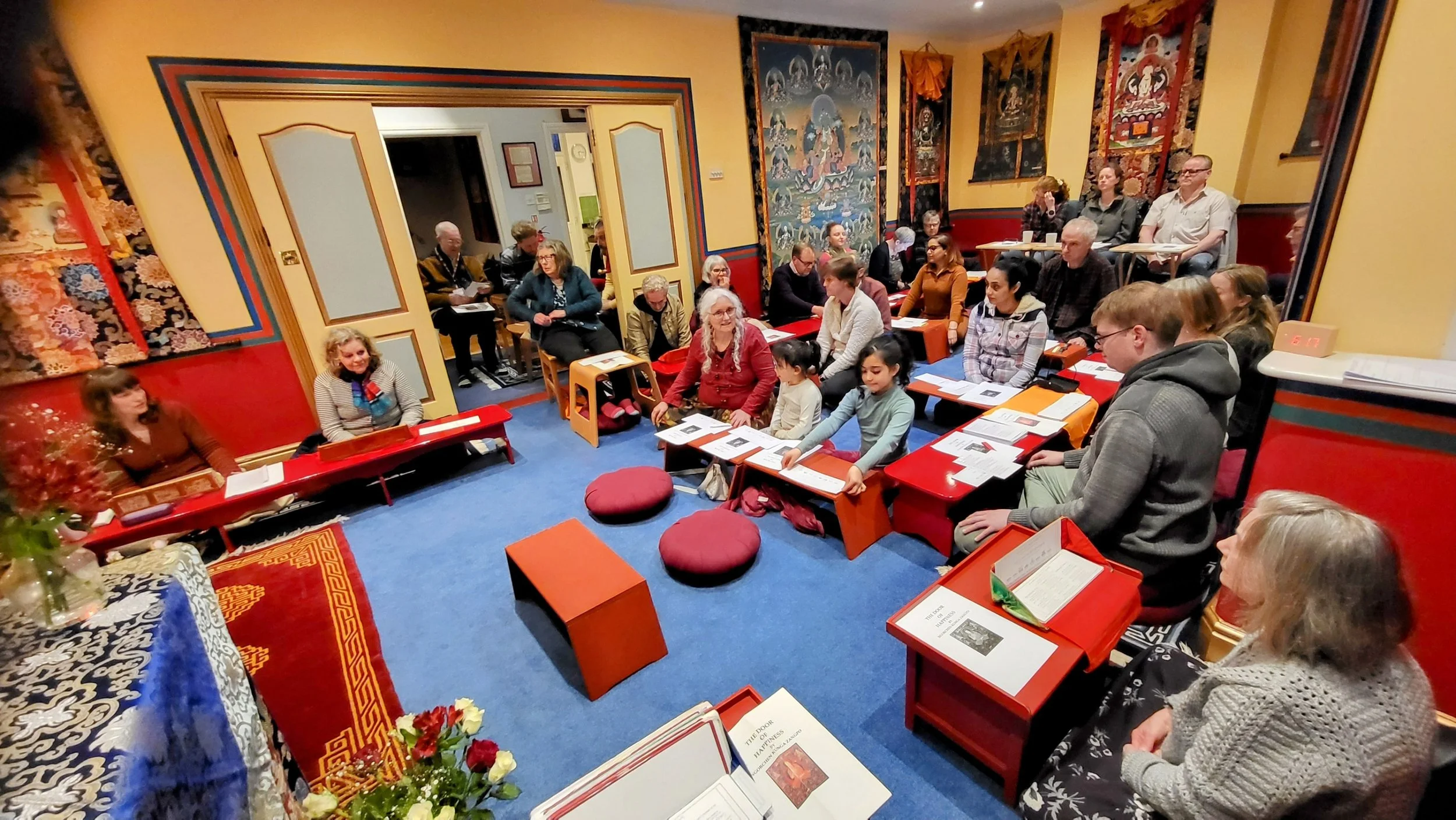 A diverse group of people, including children, adults, and elderly, are seated in a colorful, decorated room participating in a cultural or educational event. The room features vibrant wall hangings, patterned carpets, and low tables with books and papers.
